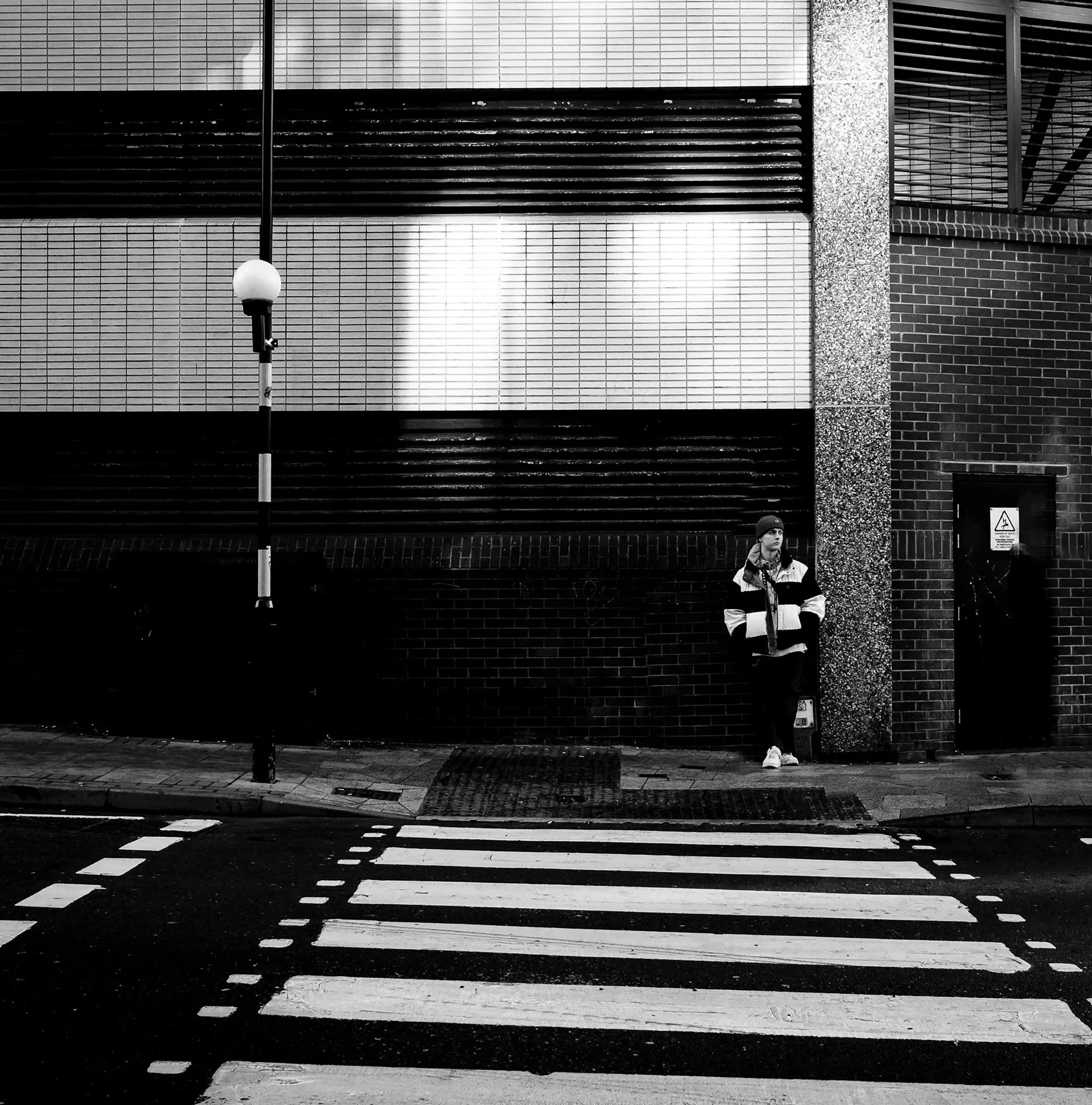 A black and white photo of a city street corner featuring a striped crosswalk, a streetlamp, and a man in a striped jacket standing near a wall.
