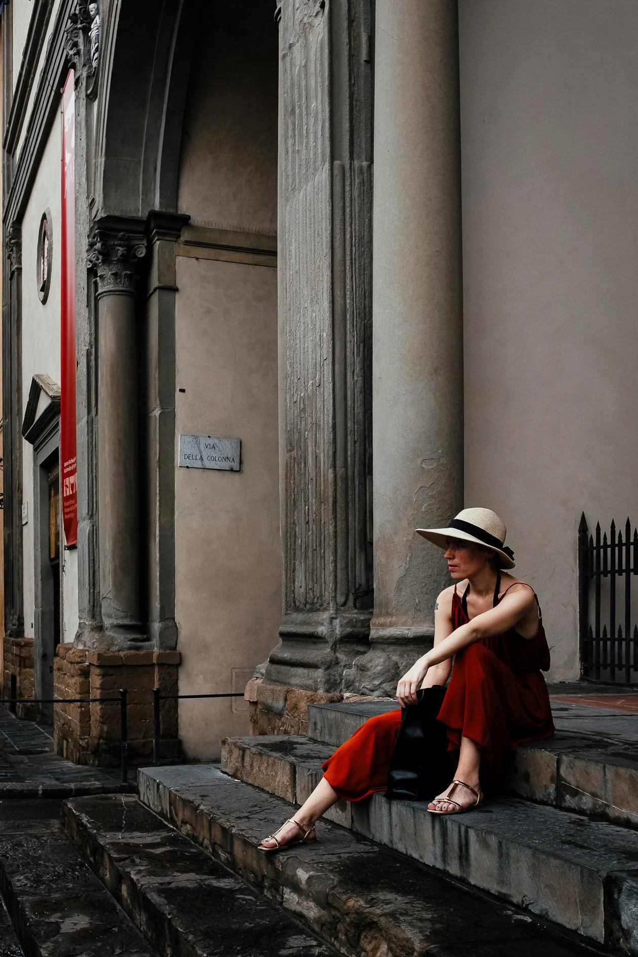 A woman in a red dress and a straw hat sits on stone steps next to a historic building with tall columns and detailed stonework in an urban setting, with a woman looking away and a street sign reading "VIA DELLA COLONNA" in the background.