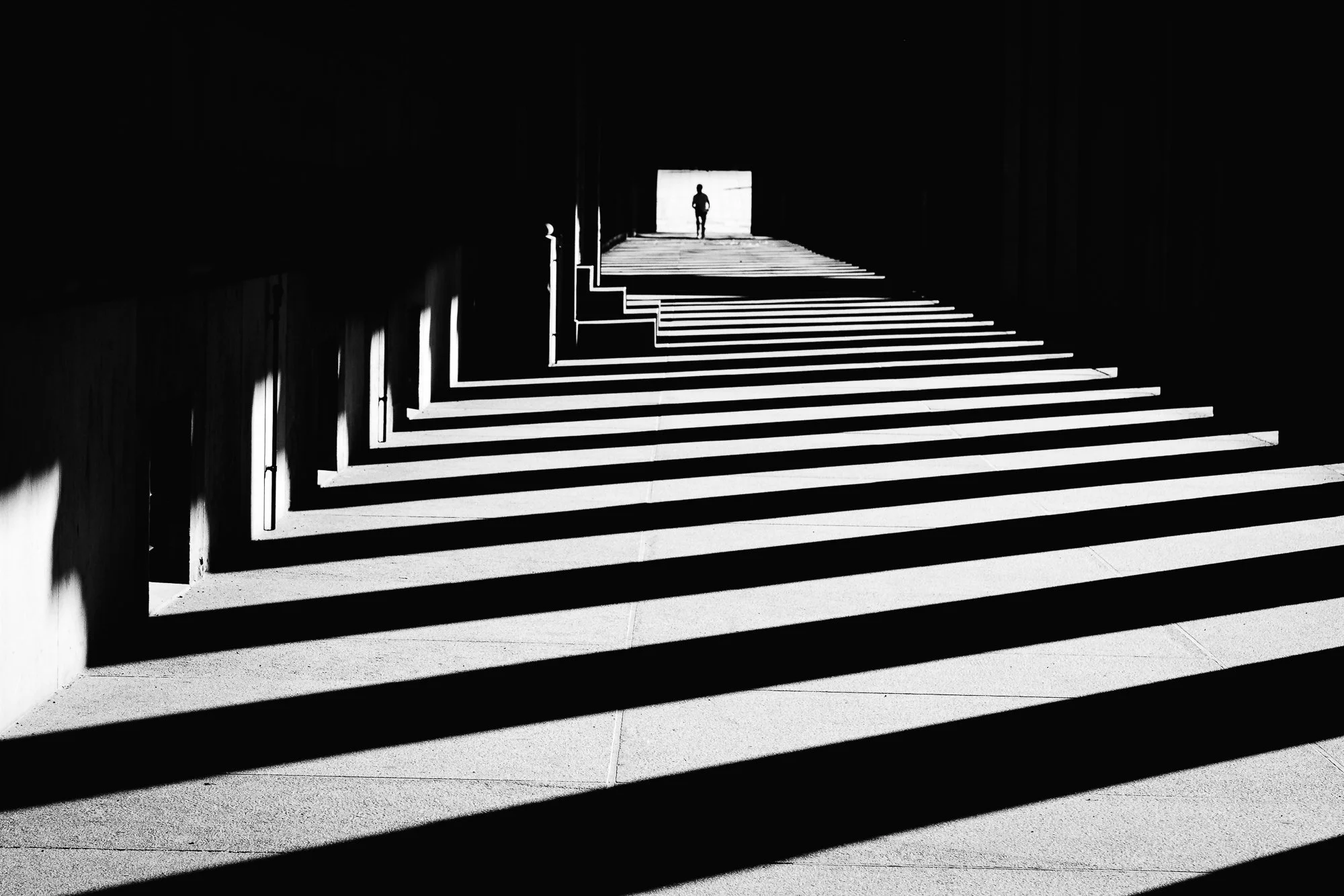 Black and white photo of staircase with strong shadows, a person walking at the top of the stairs, opening a door with bright light coming through.