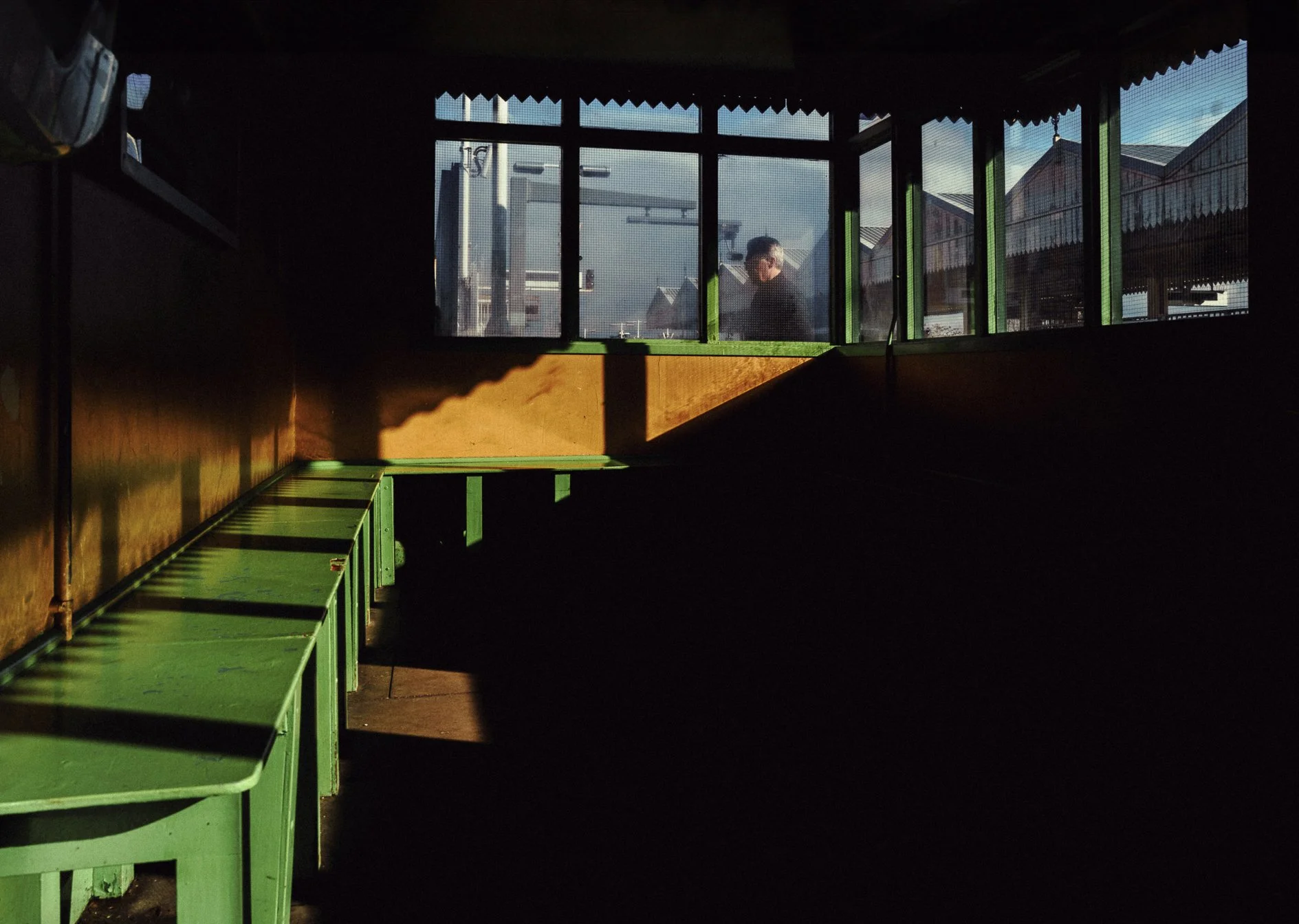 View of an indoor space with green benches along a dark wall, and large windows showing a man outside working on a structure under a blue sky.