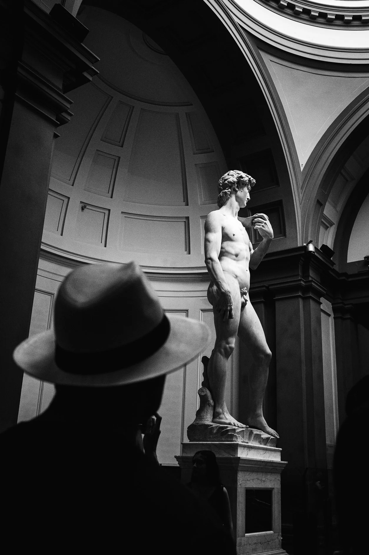 Black and white photograph of the Statue of David inside a museum, with a person in a hat observing it.