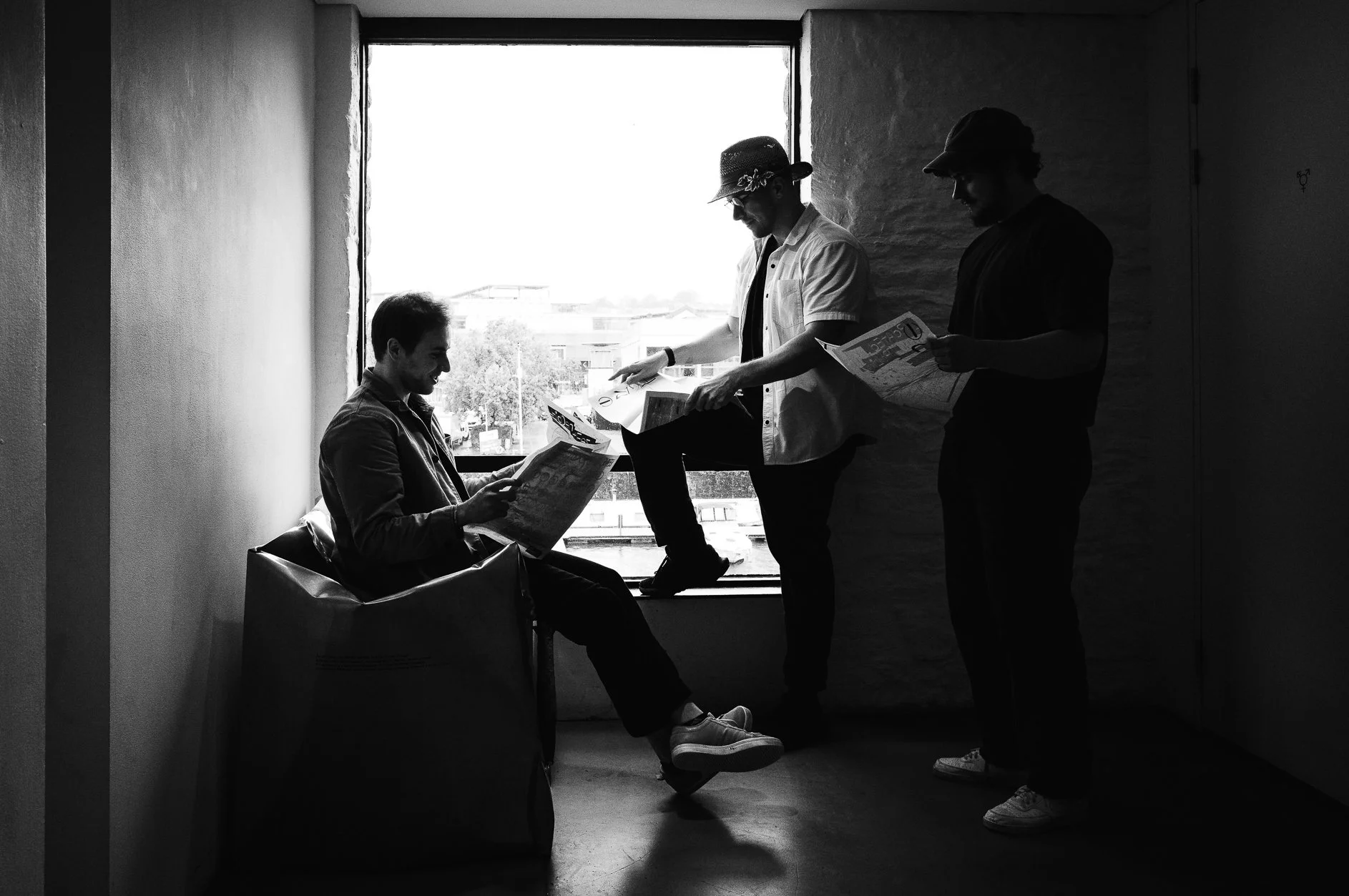 Three young men in casual clothing and hats are in a room near a large window. One man is sitting on a bench, reading a newspaper, while the other two are standing and looking at their own newspapers, engaging in conversation. The scene is in black a
