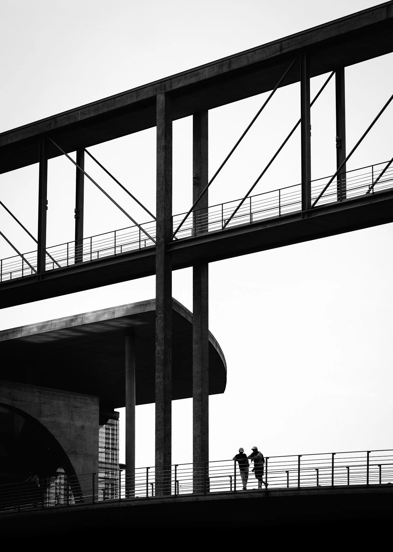 Black and white photo of a modern multi-level bridge with two people walking on the lower level, against a cloudy sky.