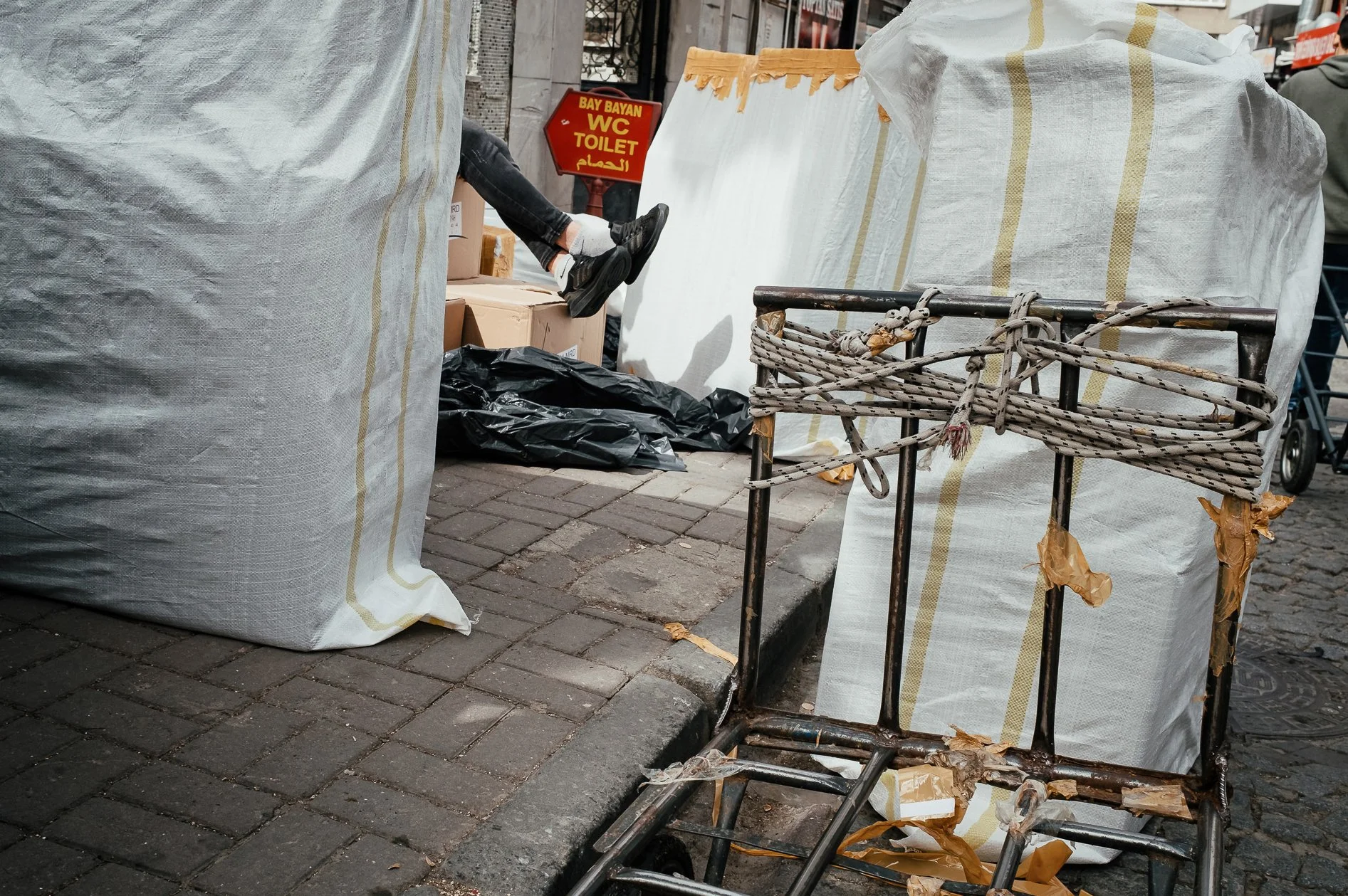 A crowded city sidewalk with large white bags, a rusted shopping cart, and a person standing near a sign that reads 'Bay Bayan WC Toilet' in English, Arabic, and Tagalog.