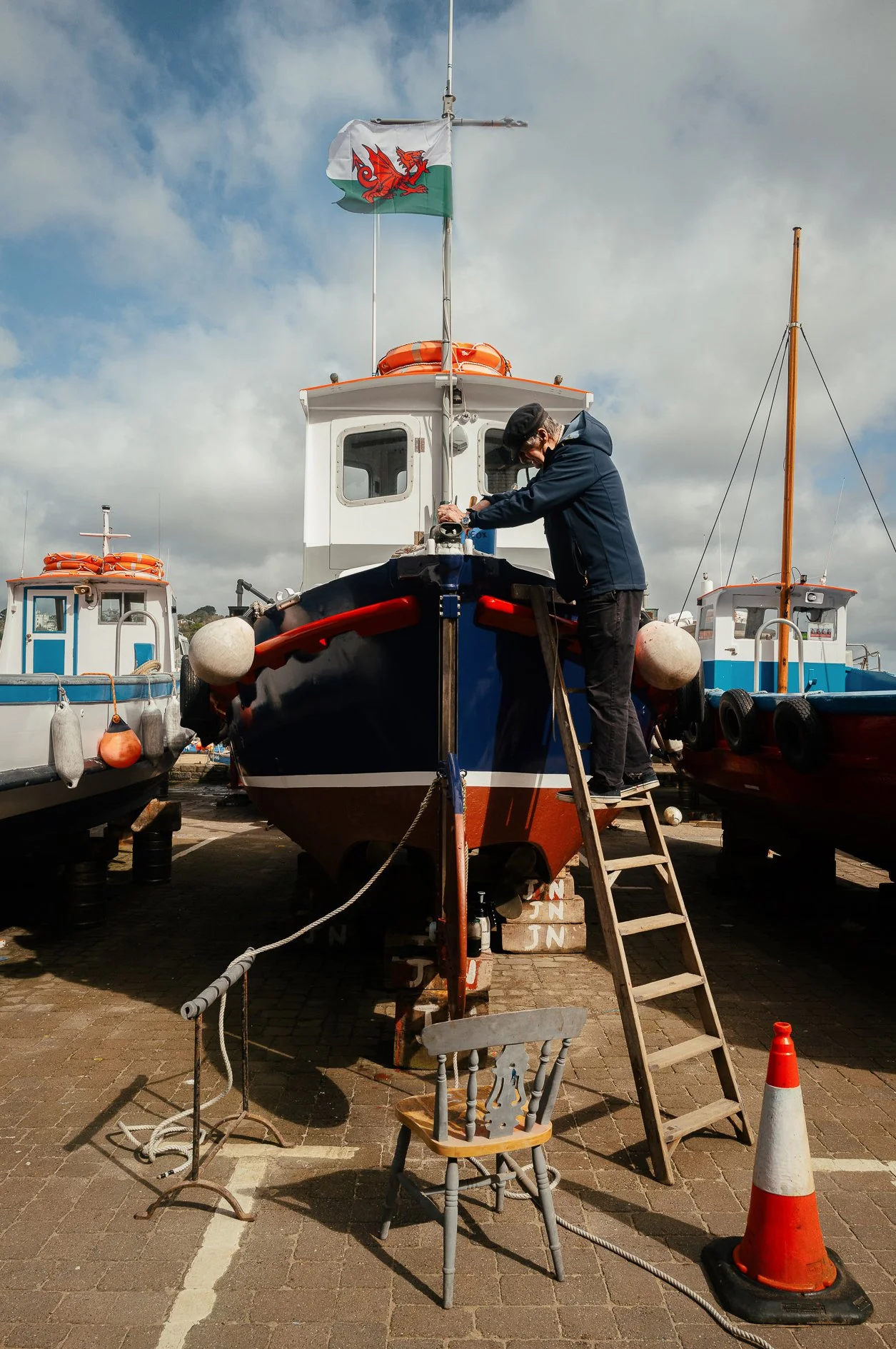 A man working on a boat in a marina, with several other boats around him. The man is standing on a ladder, painting or repairing the front part of the boat. There are orange safety cones, chairs, and ropes on the pavement. The sky is partly cloudy.