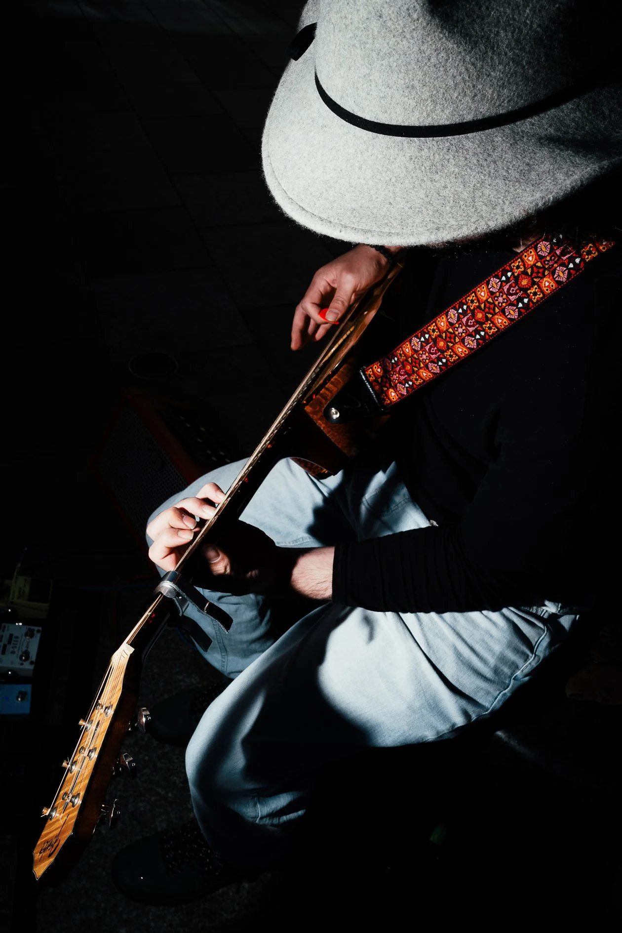 Person wearing a white hat with a black band, playing an acoustic guitar, seated with black shoes and light blue jeans, in a dark room.