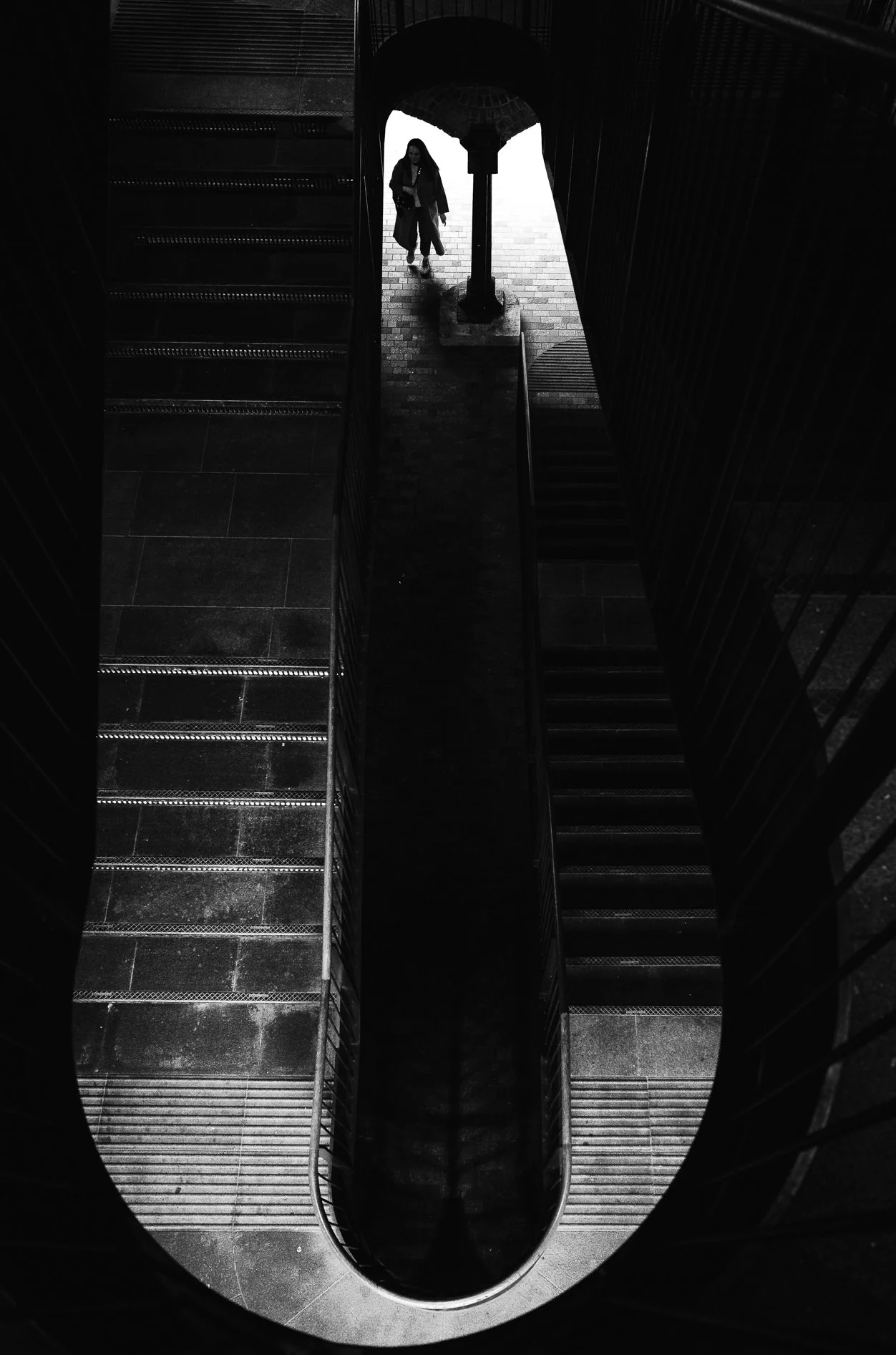 A woman walking with an umbrella down a brick corridor, viewed from above through a spiral staircase.