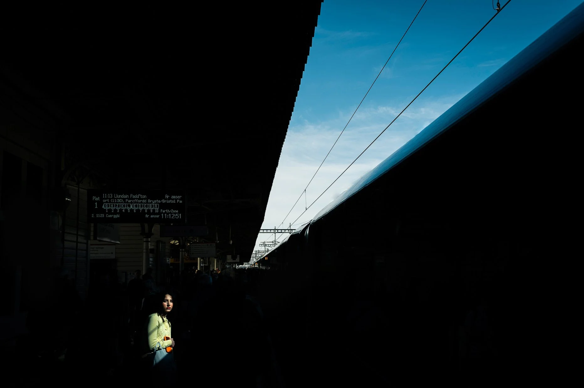 A train station platform with people waiting, a digital display board, and a train with overhead electrical lines against a blue sky.