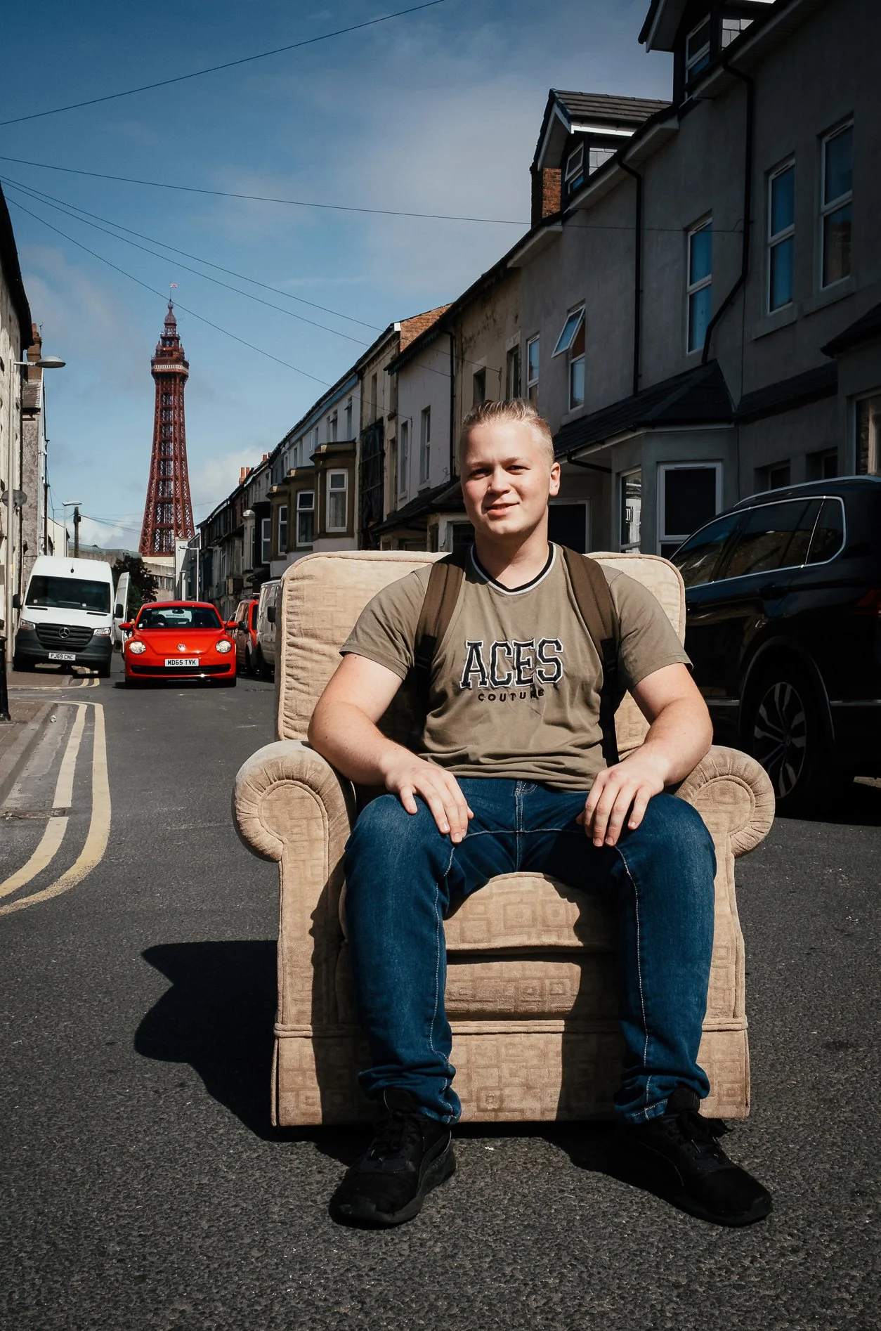 A young man sitting on a beige armchair on a city street with shops, parked cars, and the Blackpool Tower in the background.