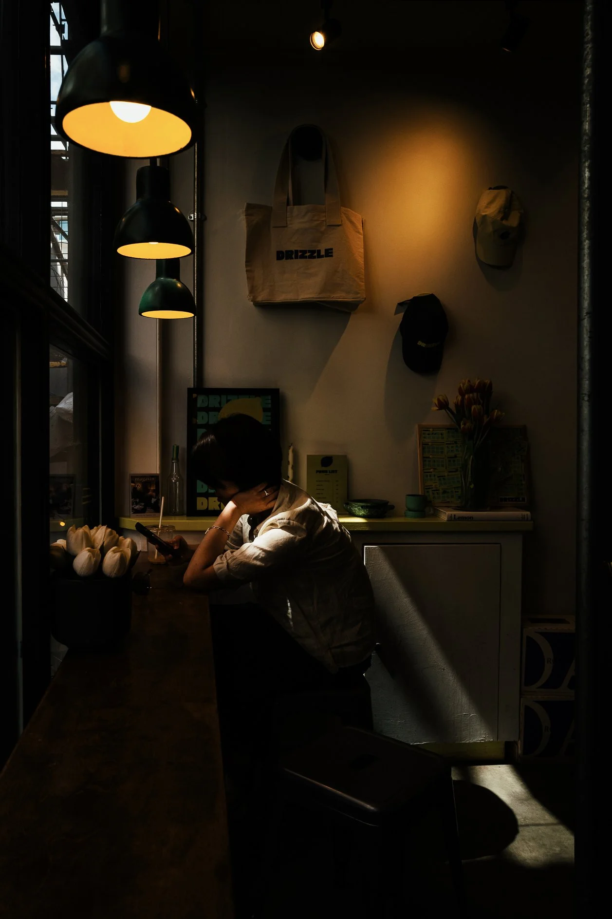 Girl sitting at a dark cafe table looking at her phone, illuminated by hanging yellow lights. Decor includes bags on the wall, a plant, and a sign in the background.