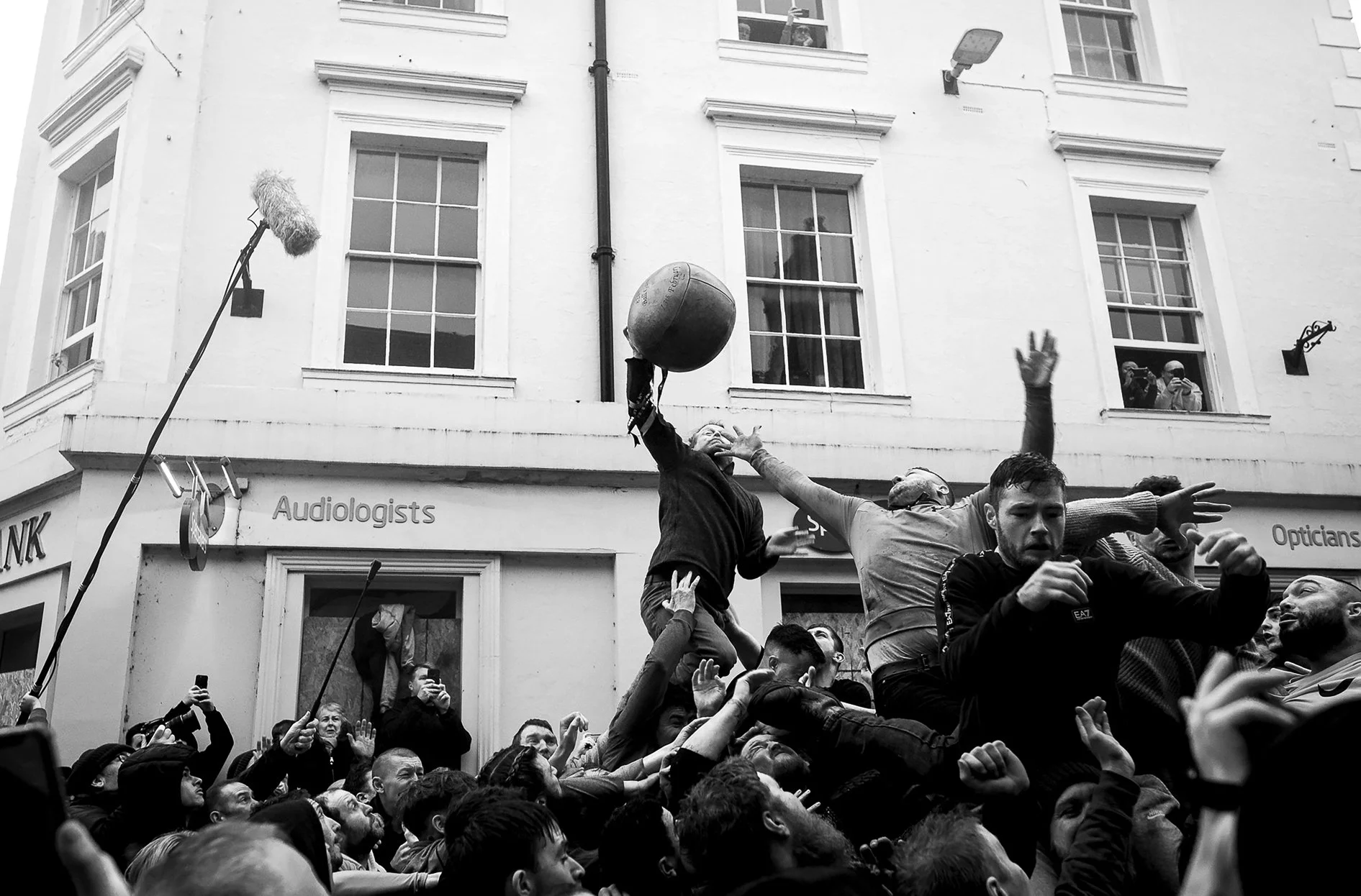 Crowd of people participating in a street event with a man raising a large ball, others reaching out or taking photos, in front of a white building with signs for audiologists and opticians.