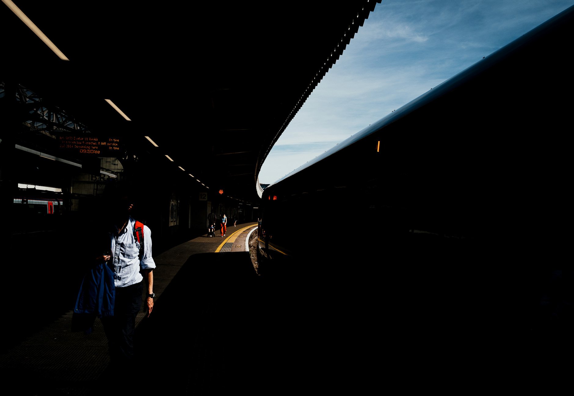 People walking on a train station platform with a curved roof, an electronic sign displaying train information, and a partly cloudy sky visible in the background.