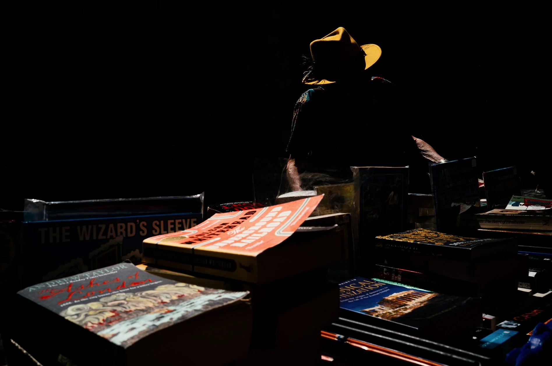 A person wearing a yellow hat stands behind a table of books in a dark room, with some books illuminated from above.