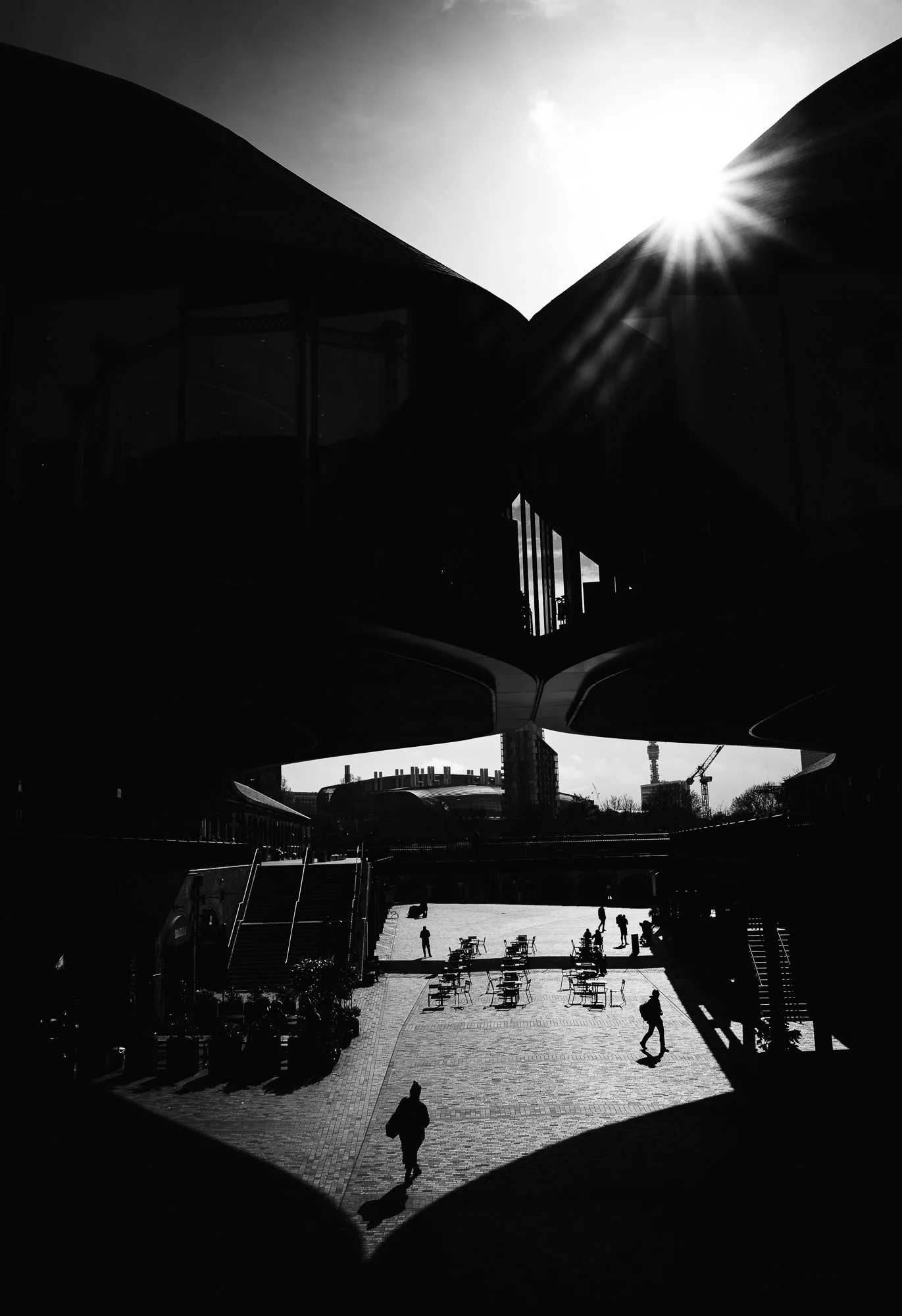 A black and white photo of a modern architectural structure with large overhanging roofs and a glass facade. The sun is shining brightly behind the building, creating lens flare and shadows. Below, people are walking and sitting in an outdoor plaza w
