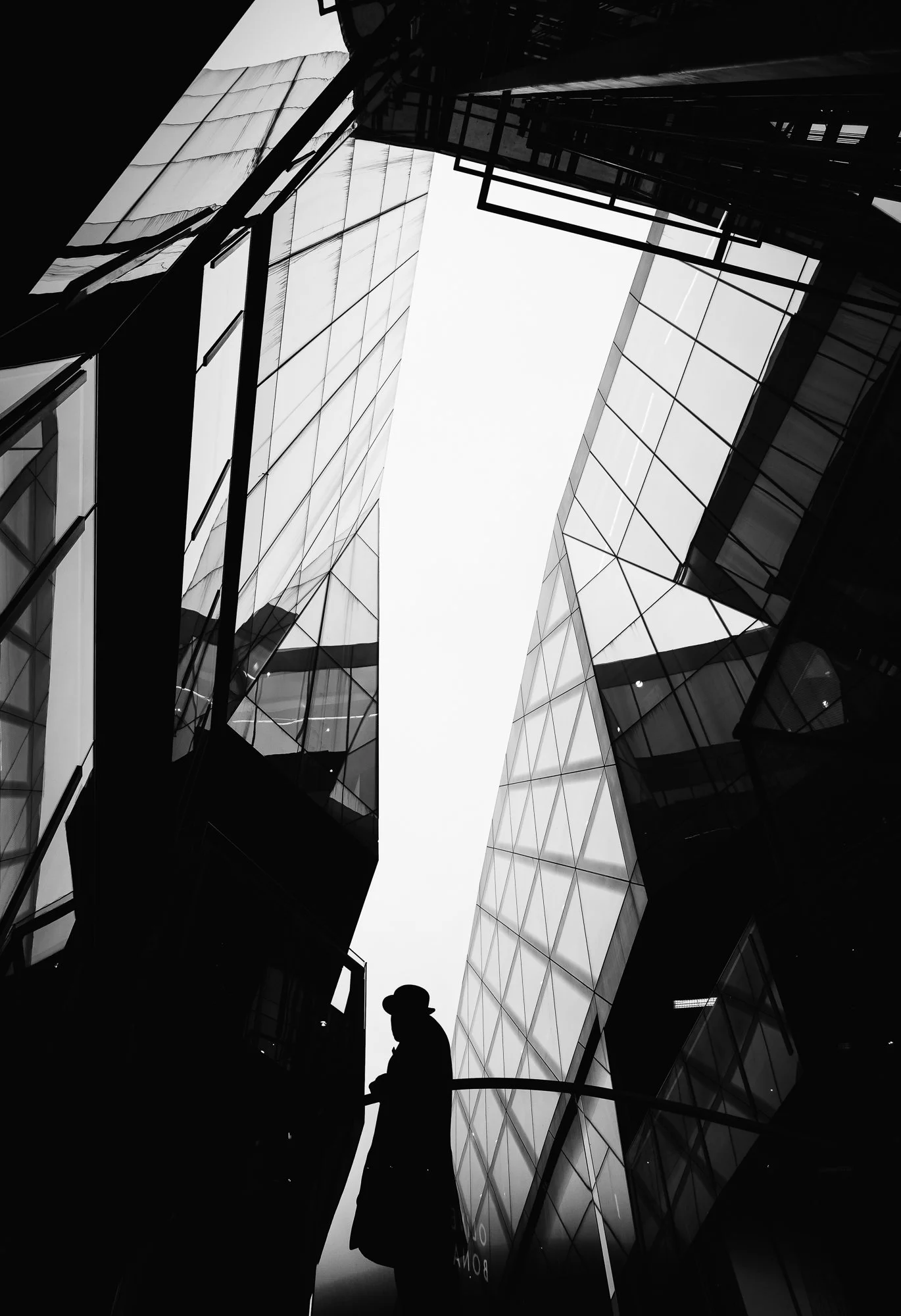 Silhouette of a person wearing a hat standing against a backdrop of modern glass buildings in an urban setting, captured from below in black and white.