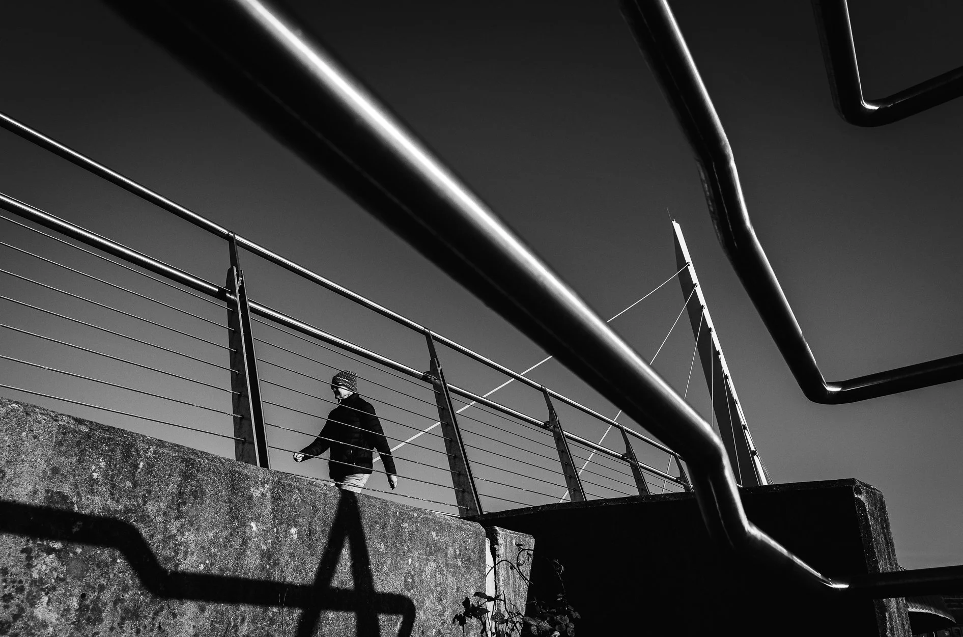 Black and white photo of a person walking along a concrete walkway with railing, with large curved metal pipes overhead and a structure in the background, under a clear sky.