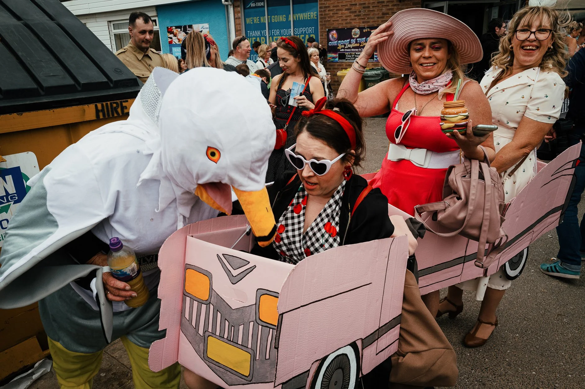 People at a street event, with some dressed in vintage or costume attire, including a woman in a large pink sunhat and a woman in a chicken costume, gathered around a pink cardboard bus prop.