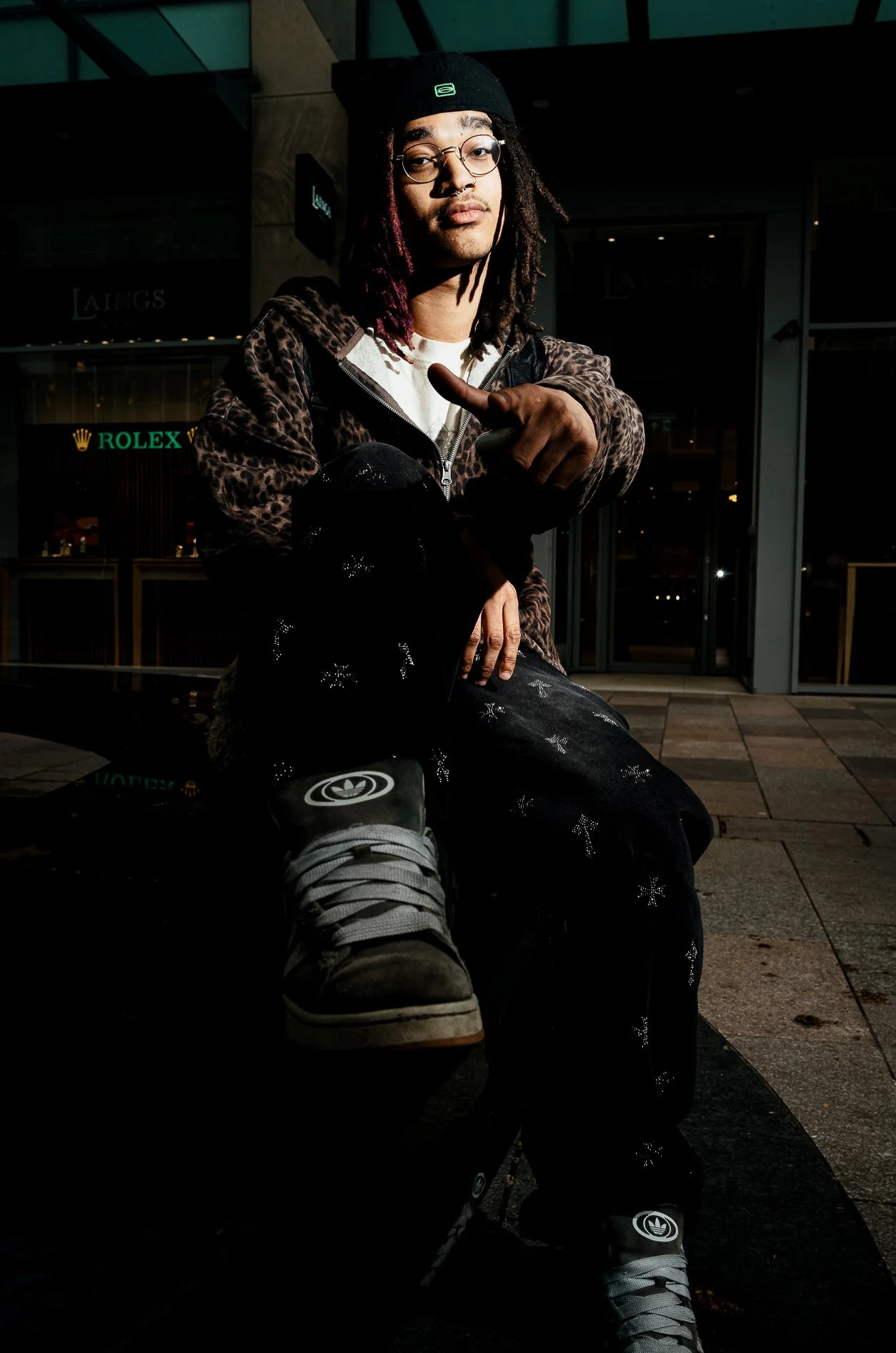 Young man with glasses and dreadlocks sitting outside at night, pointing towards the camera, wearing a black beanie, a patterned jacket, black pants with star patterns, and sneakers, with a storefront in the background.