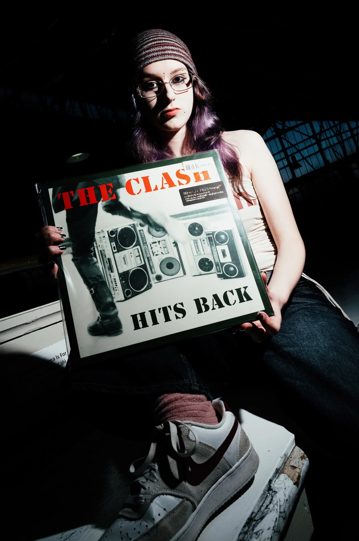Young woman with glasses and purple hair holding a vinyl record titled "The Clash Hits Back" in a dark setting with a metal building in the background.