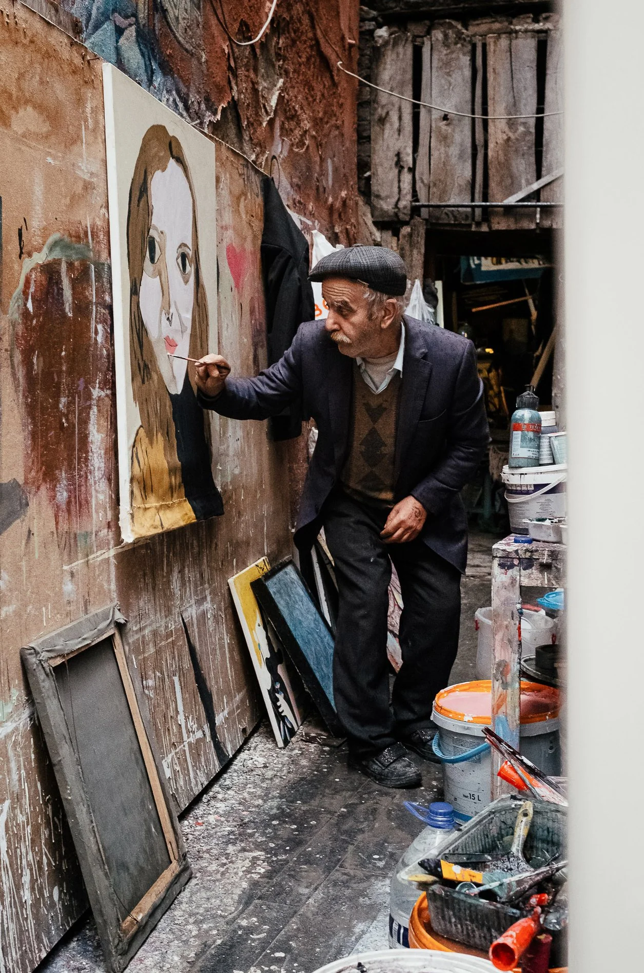 An elderly man wearing a beret and a blazer painting a portrait in a rustic art studio with dusty floors and various art supplies.