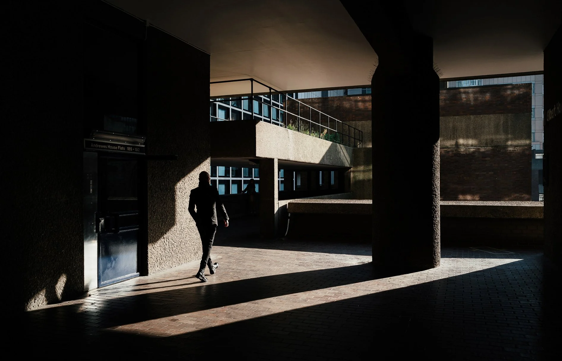 A person walking through a dark outdoor area with strong sunlight creating sharp shadows, buildings in the background, and a sign pointing to Andrews House Flats.