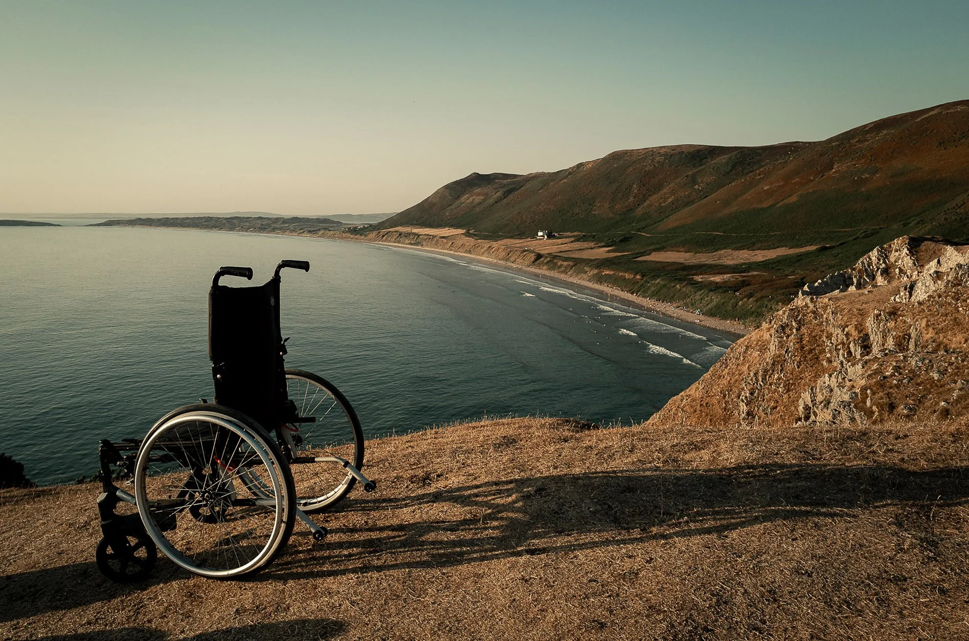 A wheelchair on a grassy cliff overlooking the ocean and rolling hills at sunset.
