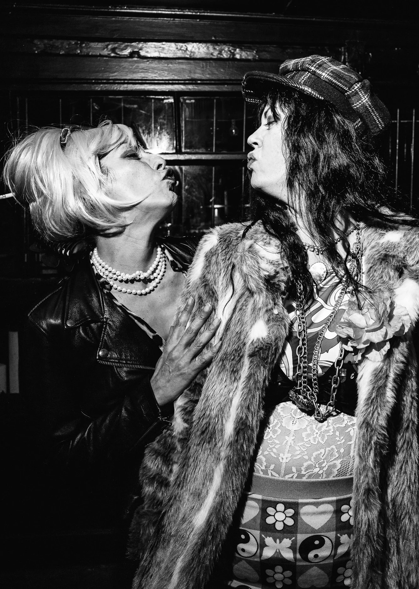 Two women facing each other close-up, one with light-colored hair, wearing a leather jacket and pearl necklaces, the other with dark hair, wearing a plaid hat and a fur coat, in black and white photo.