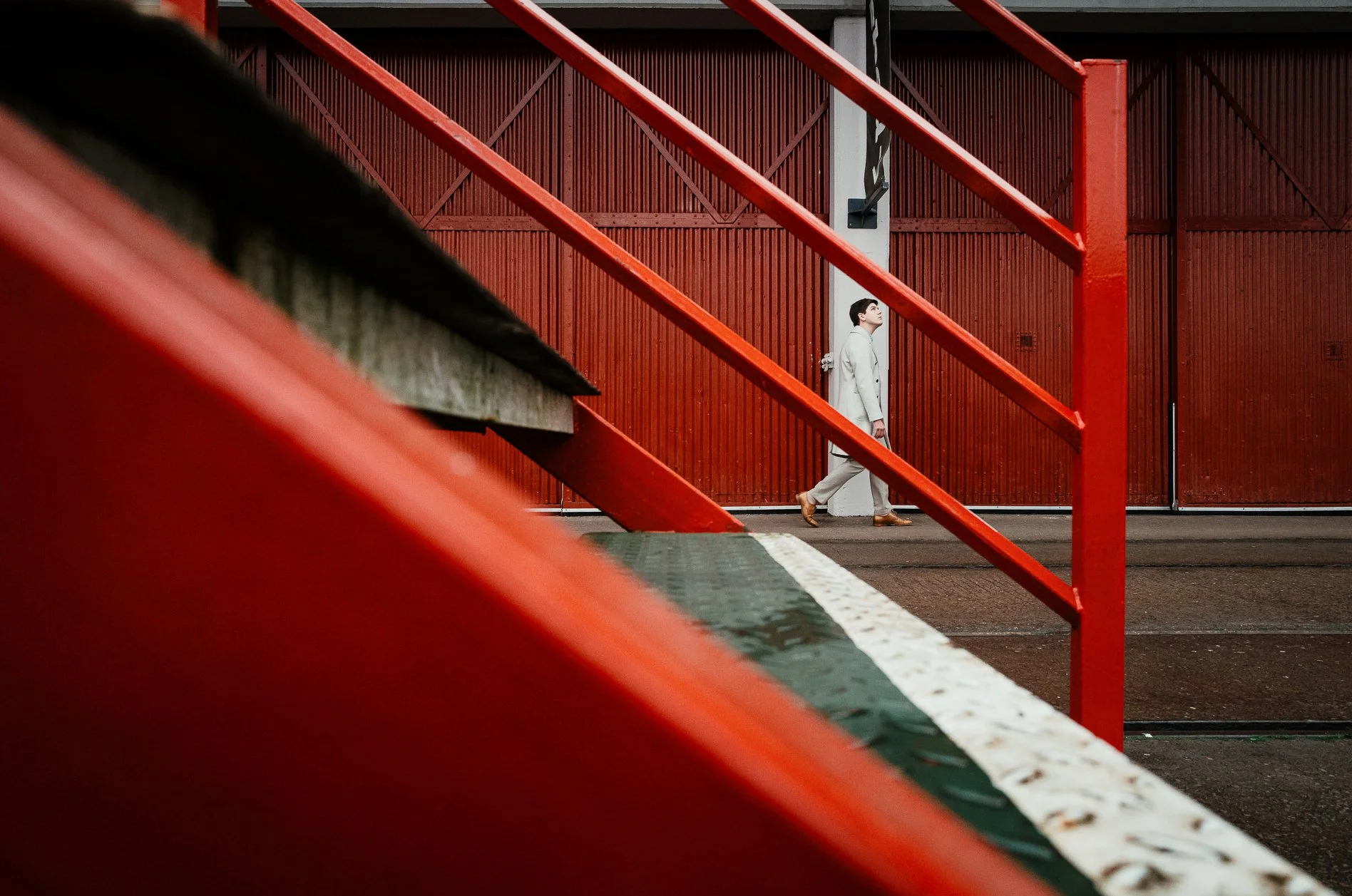 A person in a light-colored suit walking along a sidewalk next to a large red industrial building, viewed through red metal stairs and railings.