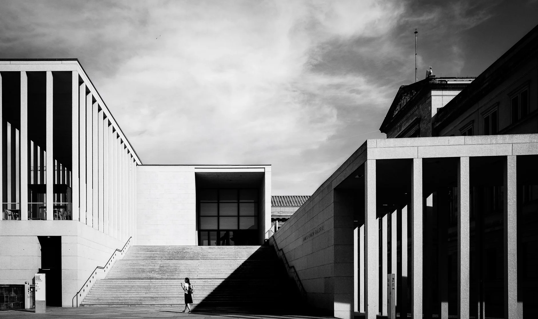 Black-and-white photo of modern building with staircase and architectural structures, a woman walking and reading a phone.