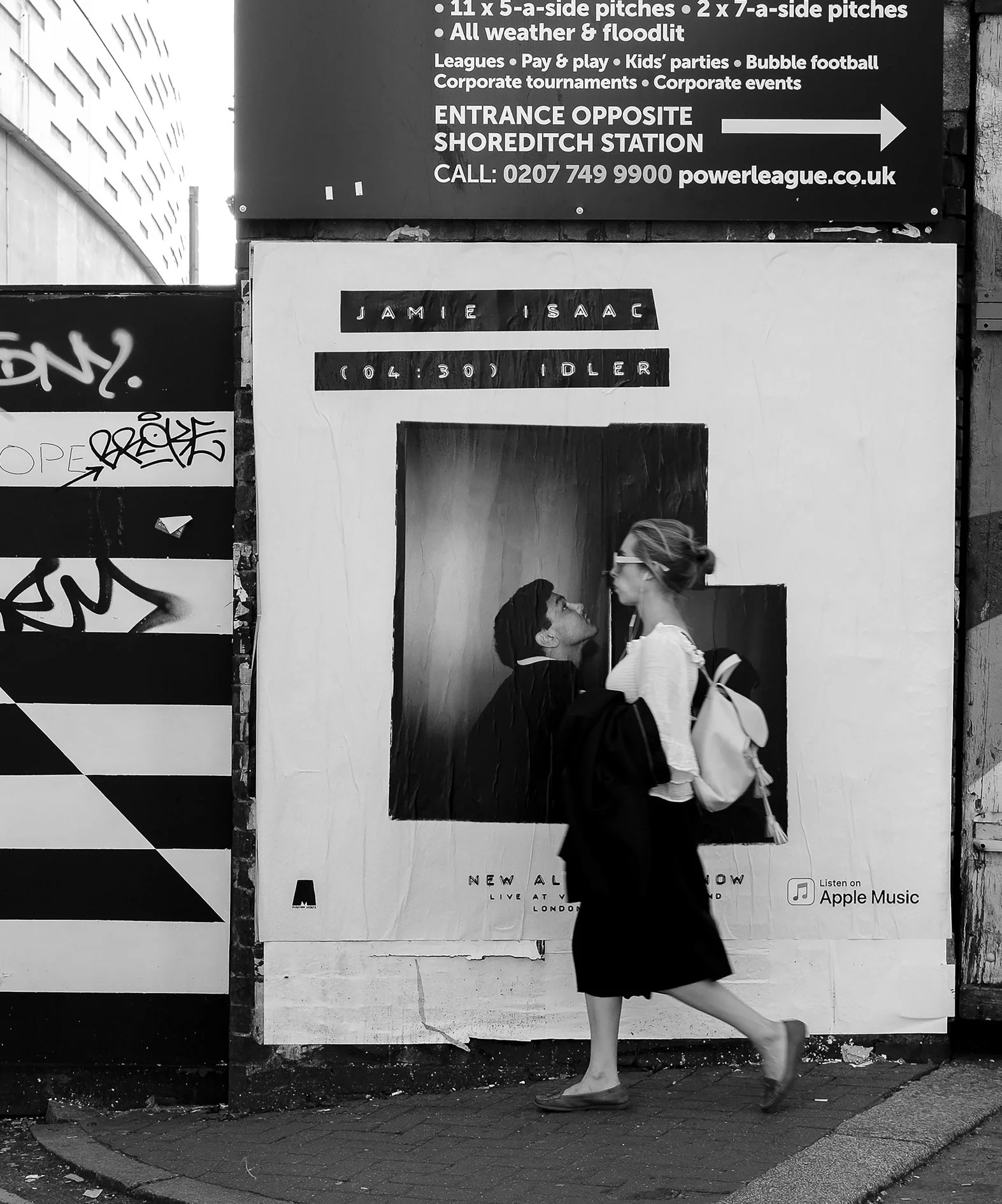 A woman crossing the street in front of a large advertisement board, with a black and white image of two women looking at each other. The woman on the street is wearing a white top, black skirt, carrying a white bag, and wearing glasses. The advertis