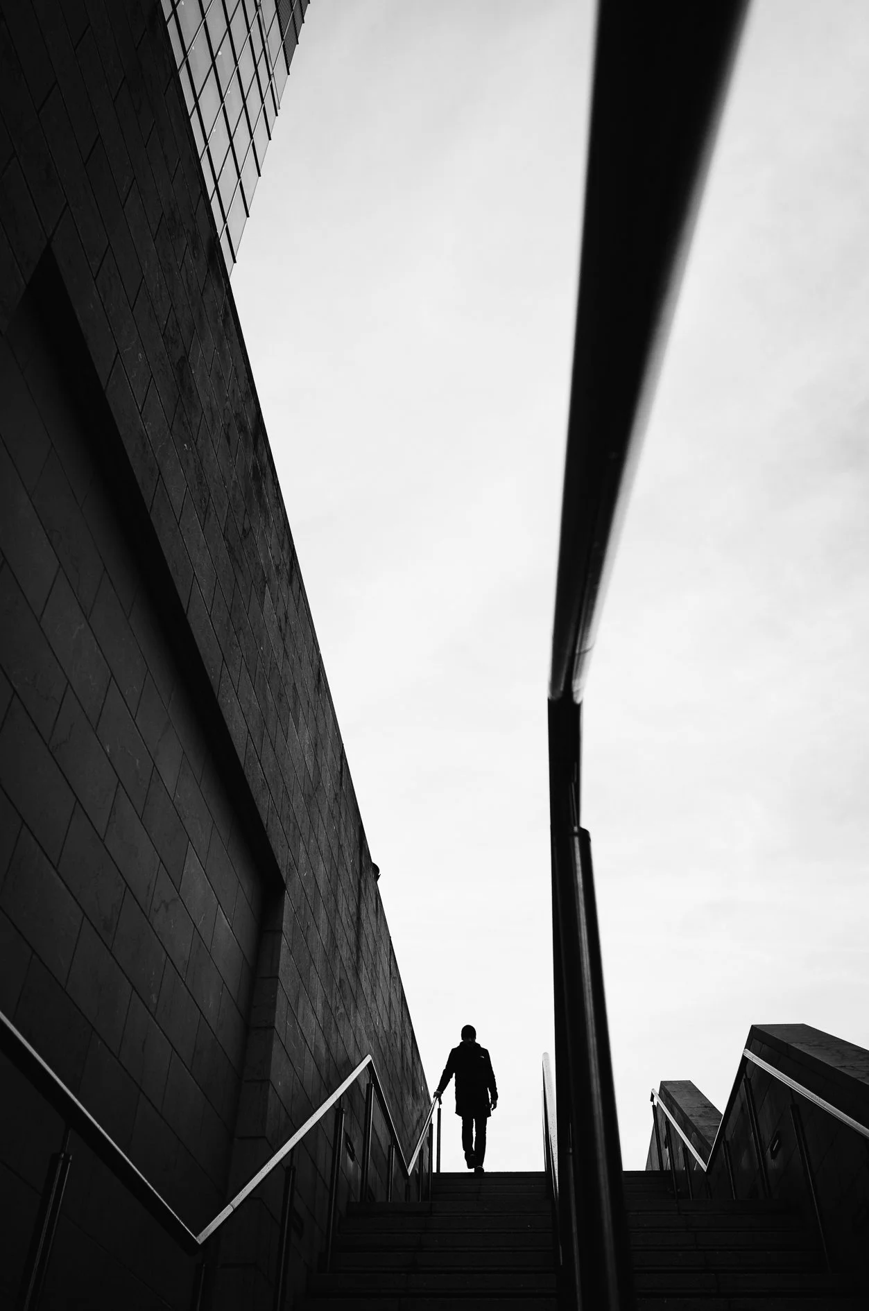 A black and white photo of a person walking up stairs outside a building with a dark brick wall. The sky is overcast and the view is from below the stairs, looking upward.