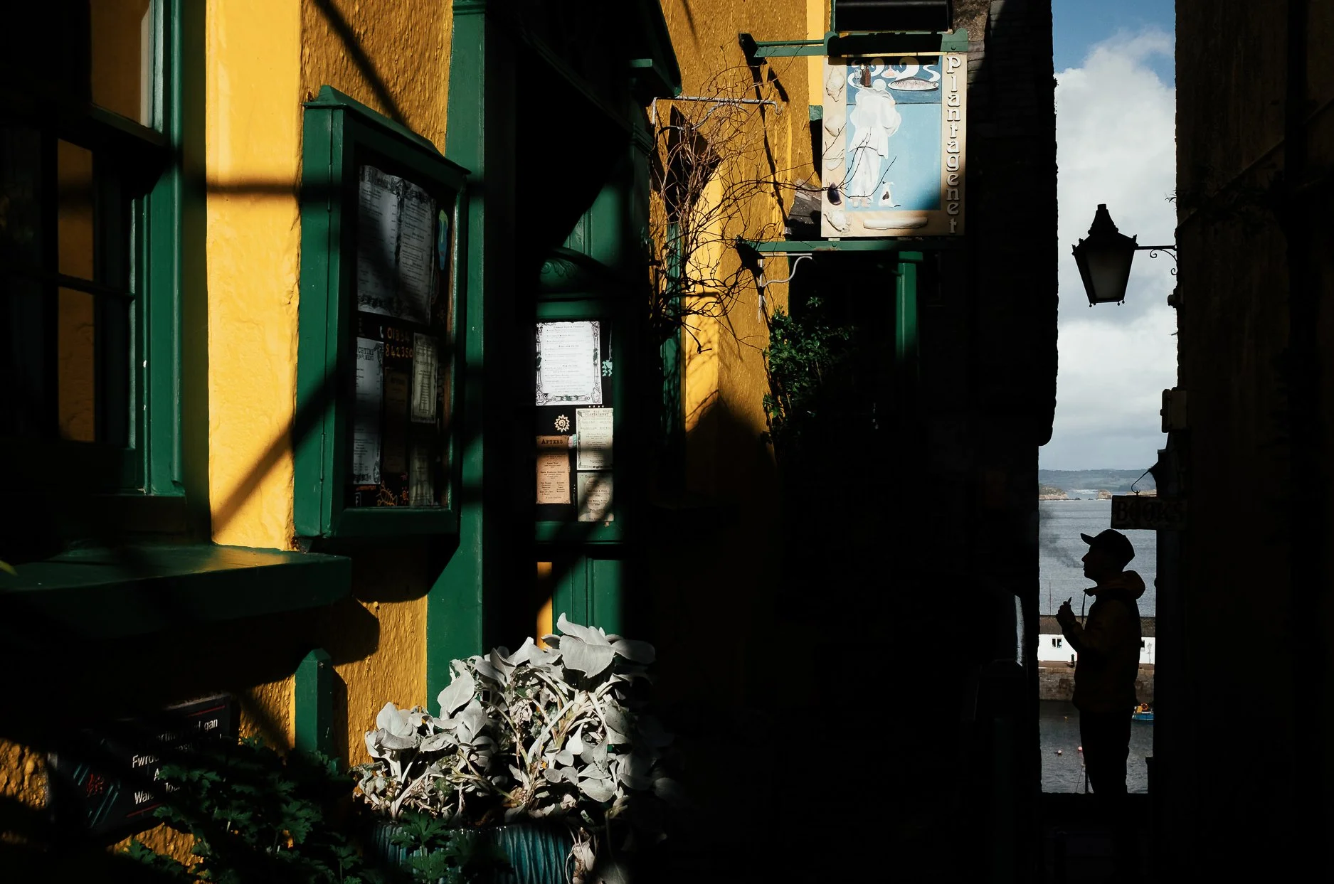Narrow alleyway with yellow walls and green window frames, shadow from a street sign, a person with a hat holding a phone or camera, standing near a view of the water and sky in the background.