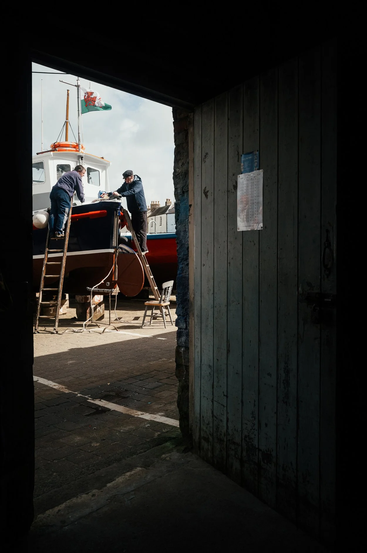 Two men working on a boat in a boatyard as seen through a dark doorway; one man stands on a ladder, and the other leans over the boat, which is elevated on supports; a Welsh flag is flying on the boat.