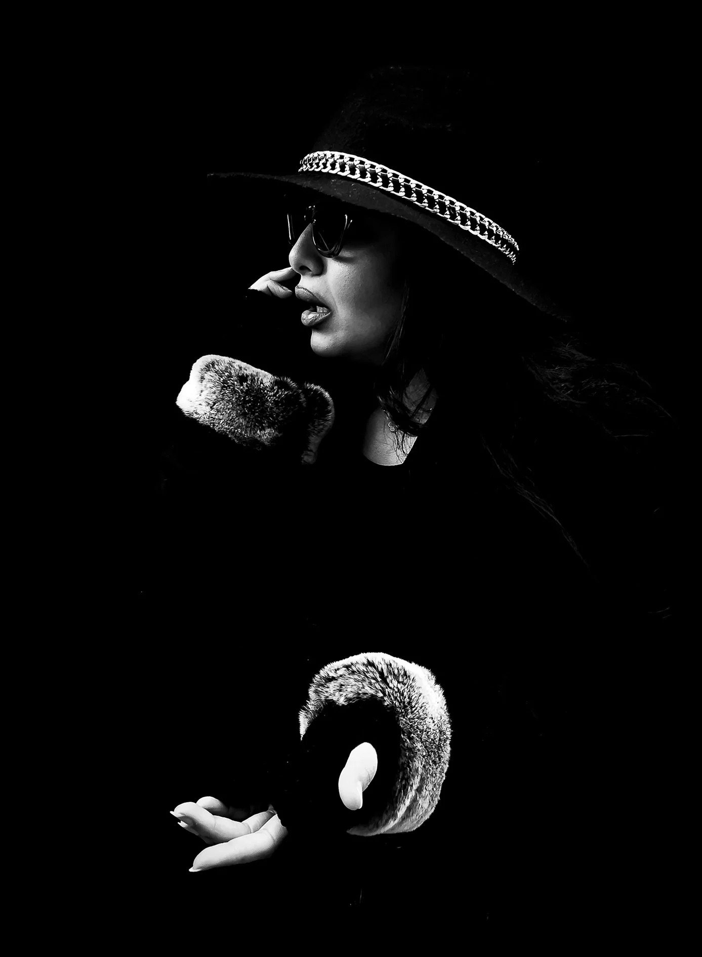 A black and white photo of a woman wearing a large hat with a chain embellishment, big sunglasses, and holding a faux fur stole, with a reflection of a swan in her hand.