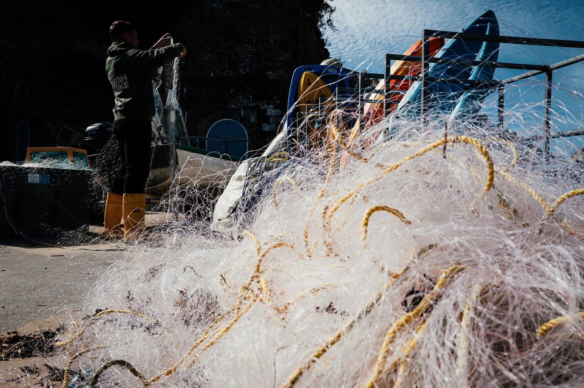 A person standing near fishing nets and equipment, with colorful kayaks in the background and an overcast sky.