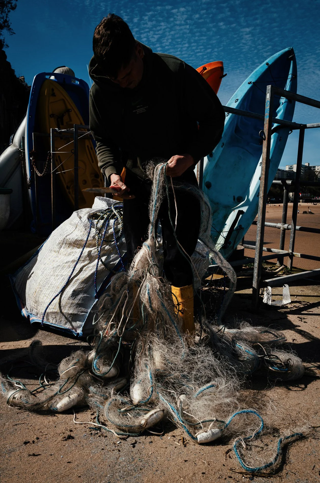 A person on a beach repairs or sorts through tangled fishing nets and ropes, with kayaks and a metal railing in the background under a blue sky.