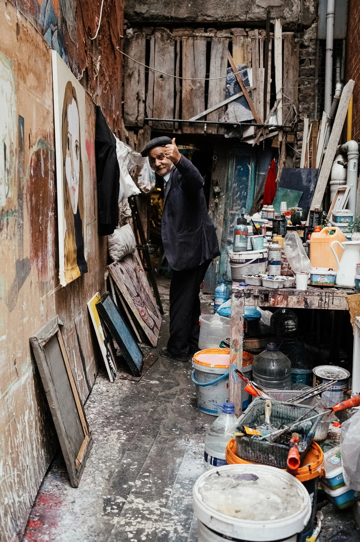 An older man in a dark jacket and beret giving a thumbs up and smiling inside an artist's studio cluttered with paintings, paint cans, brushes, and art supplies.