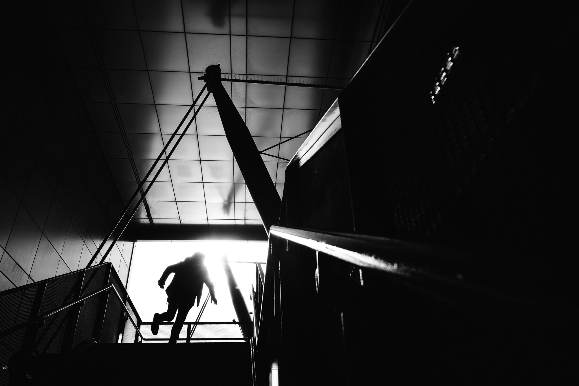 A silhouette of a person walking down stairs with sunlight in the background, in a modern building with tiled ceiling and metal railings.