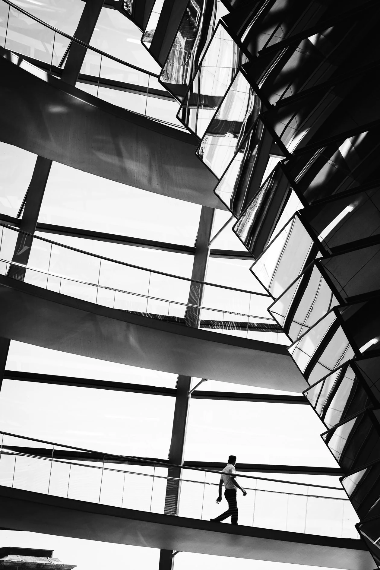 Black and white photo of a modern glass building interior with multiple floors and walkways. A person is walking across one of the upper floors.