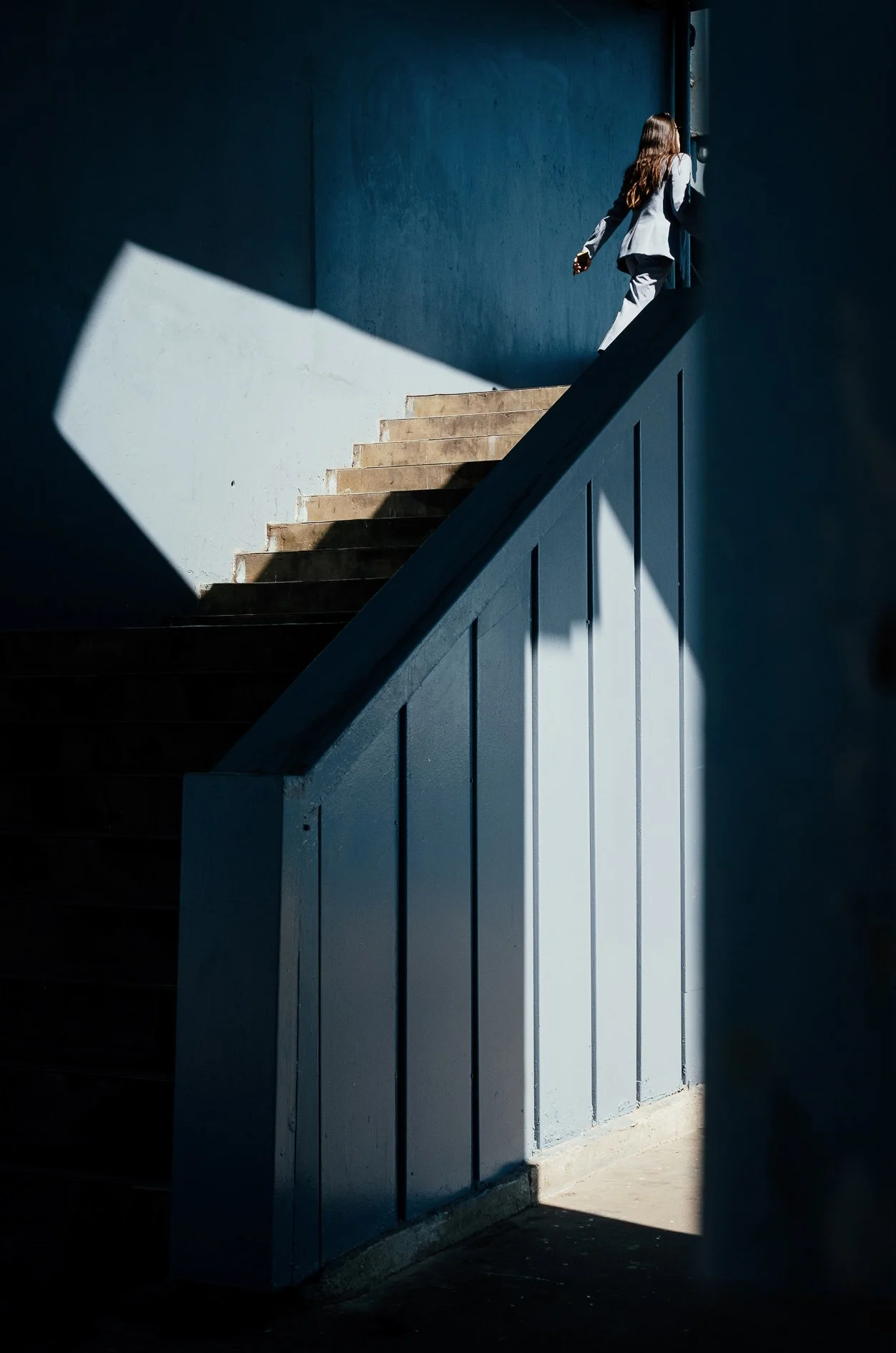 A person in a gray suit walking up a staircase in a building with blue walls, partially illuminated by sunlight casting shadows on the stairs and wall.