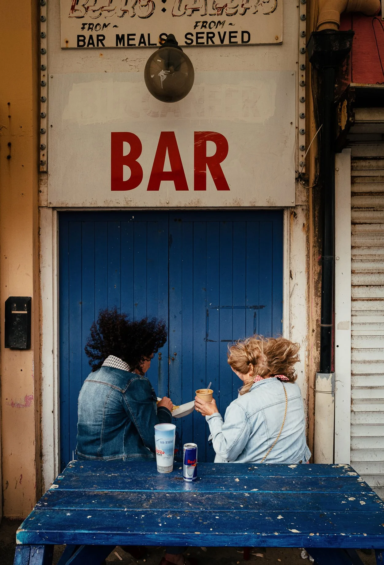 Two women eating outside a bar, standing in front of a blue door with a sign that reads 'BAR'. One woman has curly black hair and is wearing a denim jacket, the other has light brown hair tied back and is also wearing a denim jacket. There are drinks
