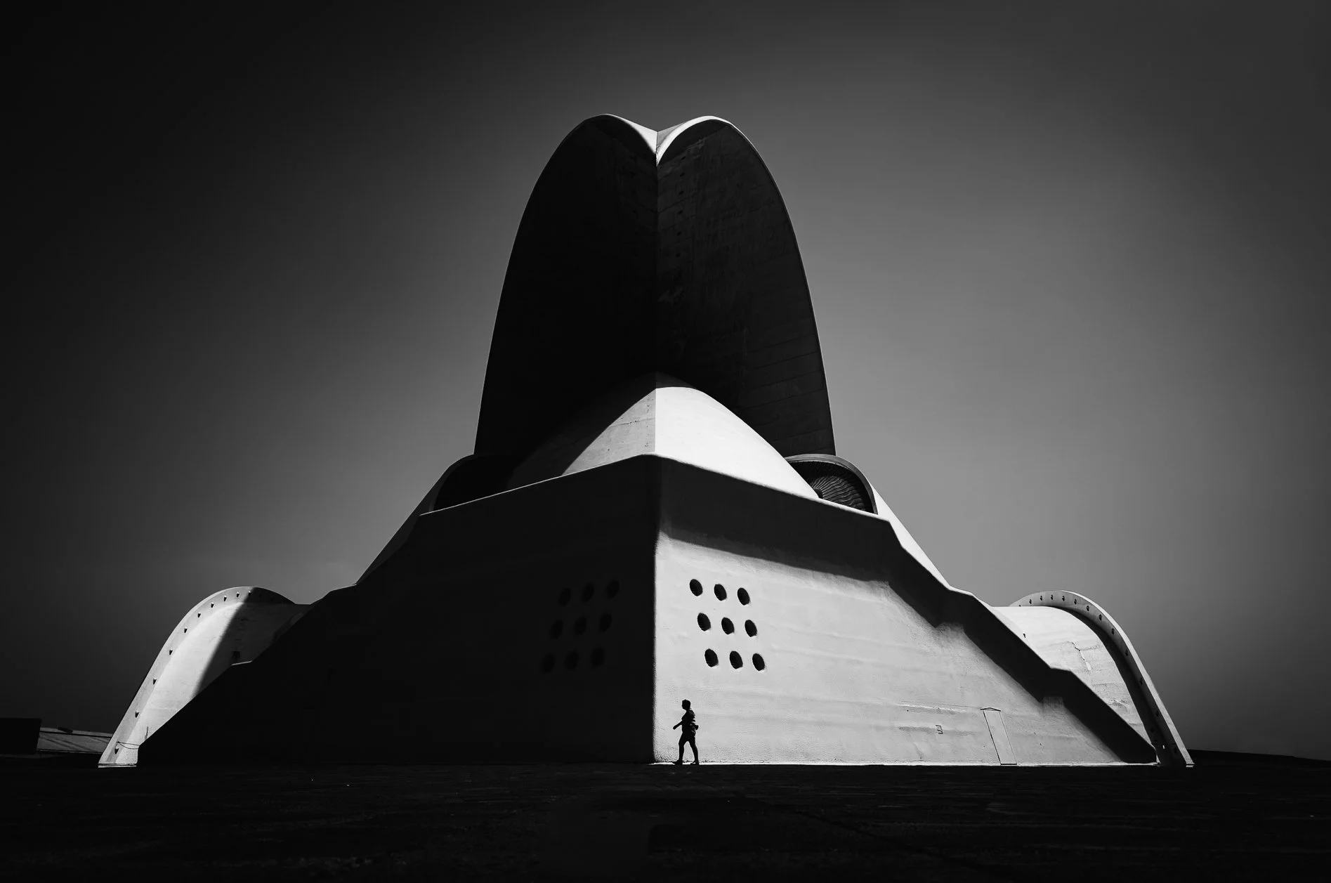 Black and white photo of a large futuristic building with a person standing near the base, showcasing the scale of the structure. The building has smooth curves and bold geometric shapes, with a clear sky background.