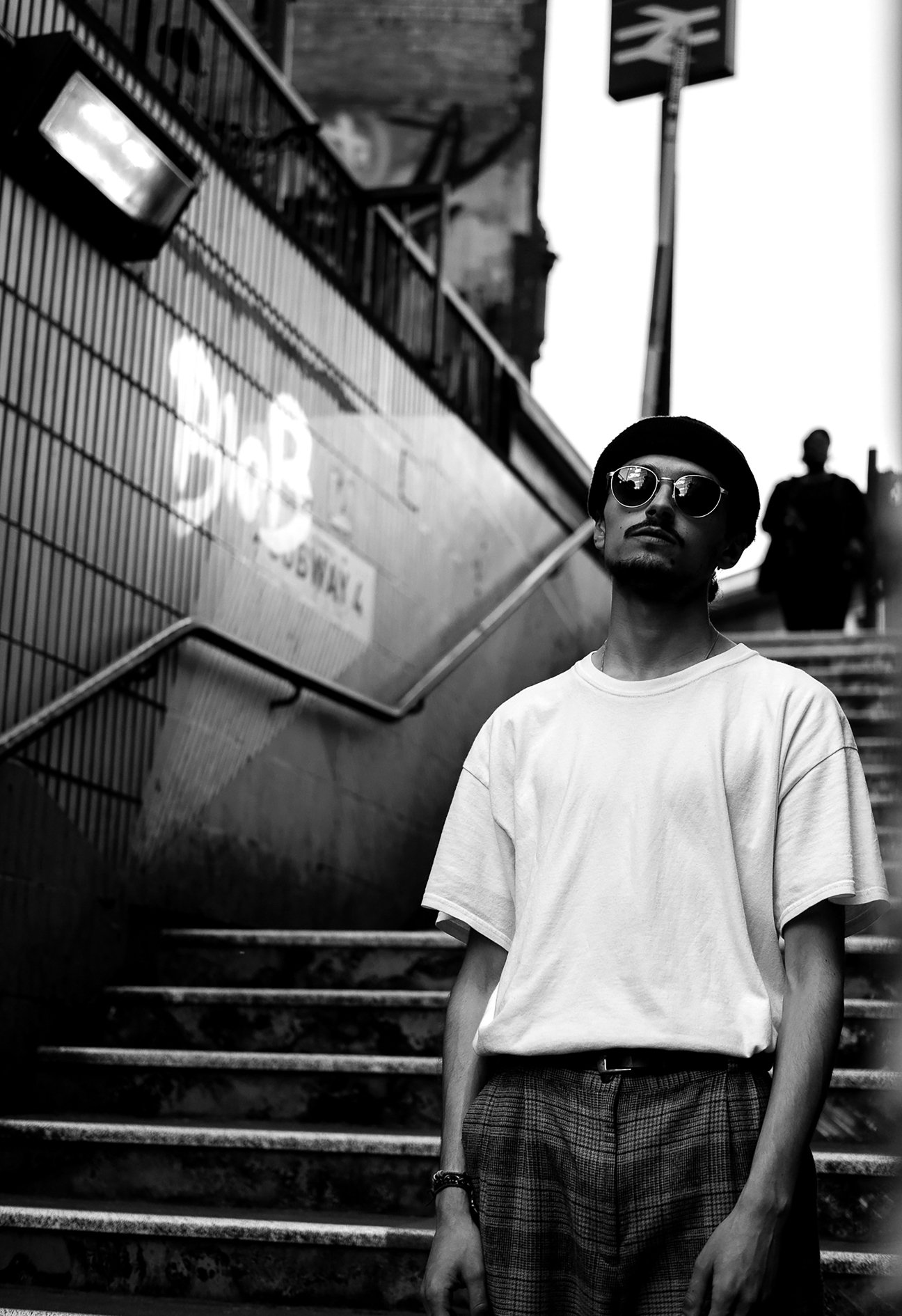 A young man with dark sunglasses, a black hat, a white t-shirt, and plaid pants standing on stairs outside a building with a graffiti-covered wall and a sign in the background.