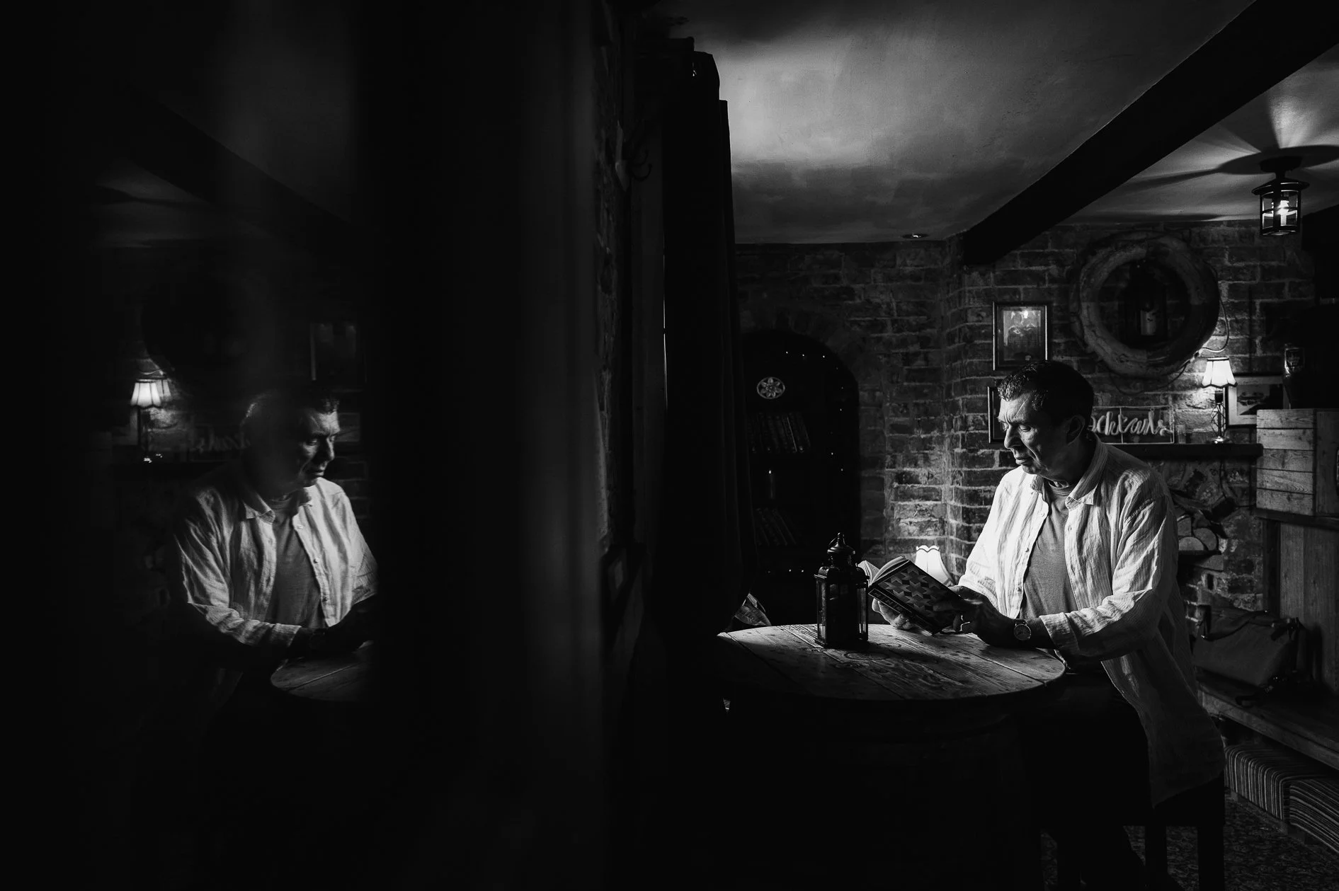 A black and white photo of a man reading a book at a rustic wooden table in a cozy room with brick walls, a mirror, and lamps.