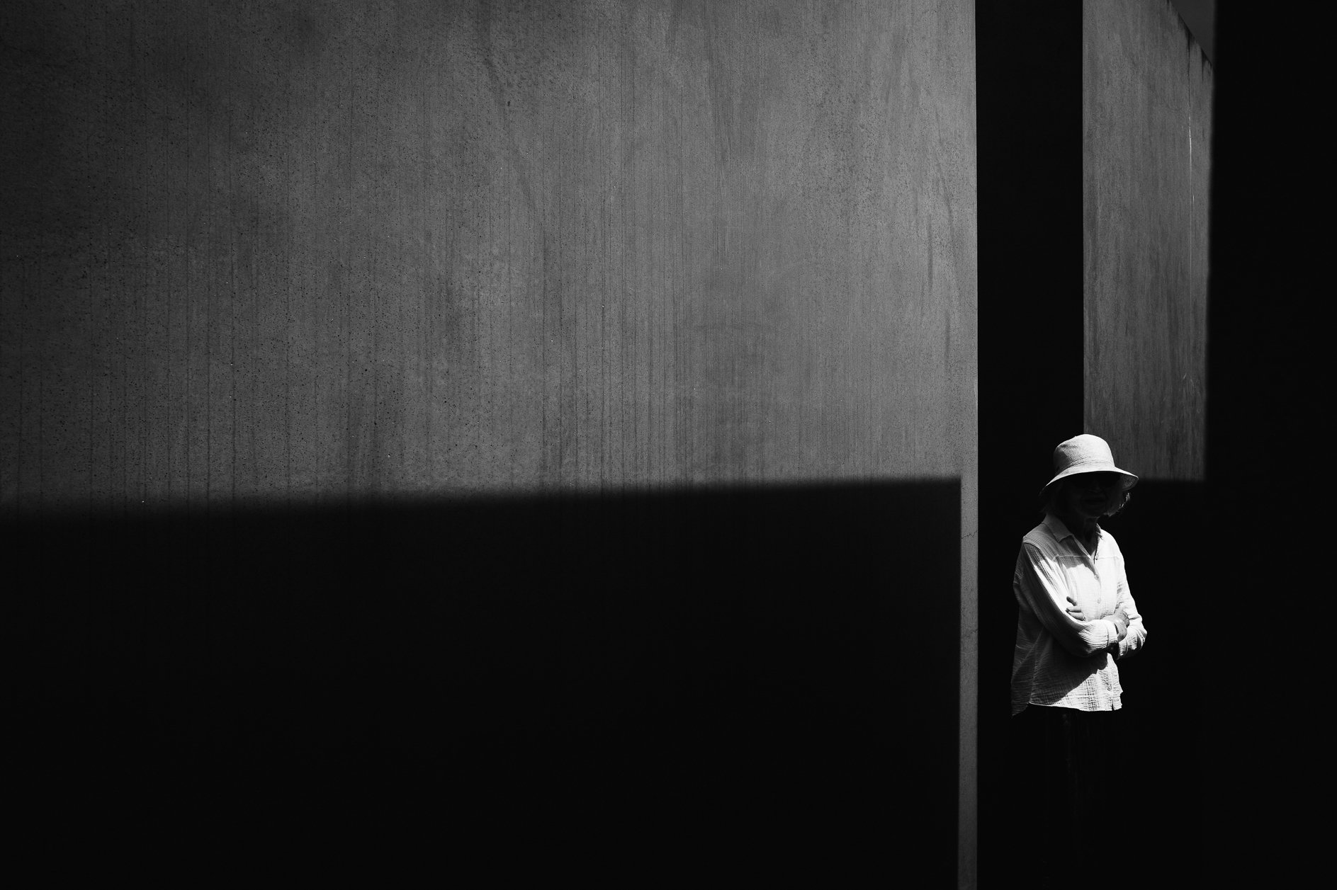 A black and white photo of a woman standing in shadow, wearing a wide-brimmed hat and a light-colored shirt, with her arms crossed, positioned near a bright wall with geometric shadows.