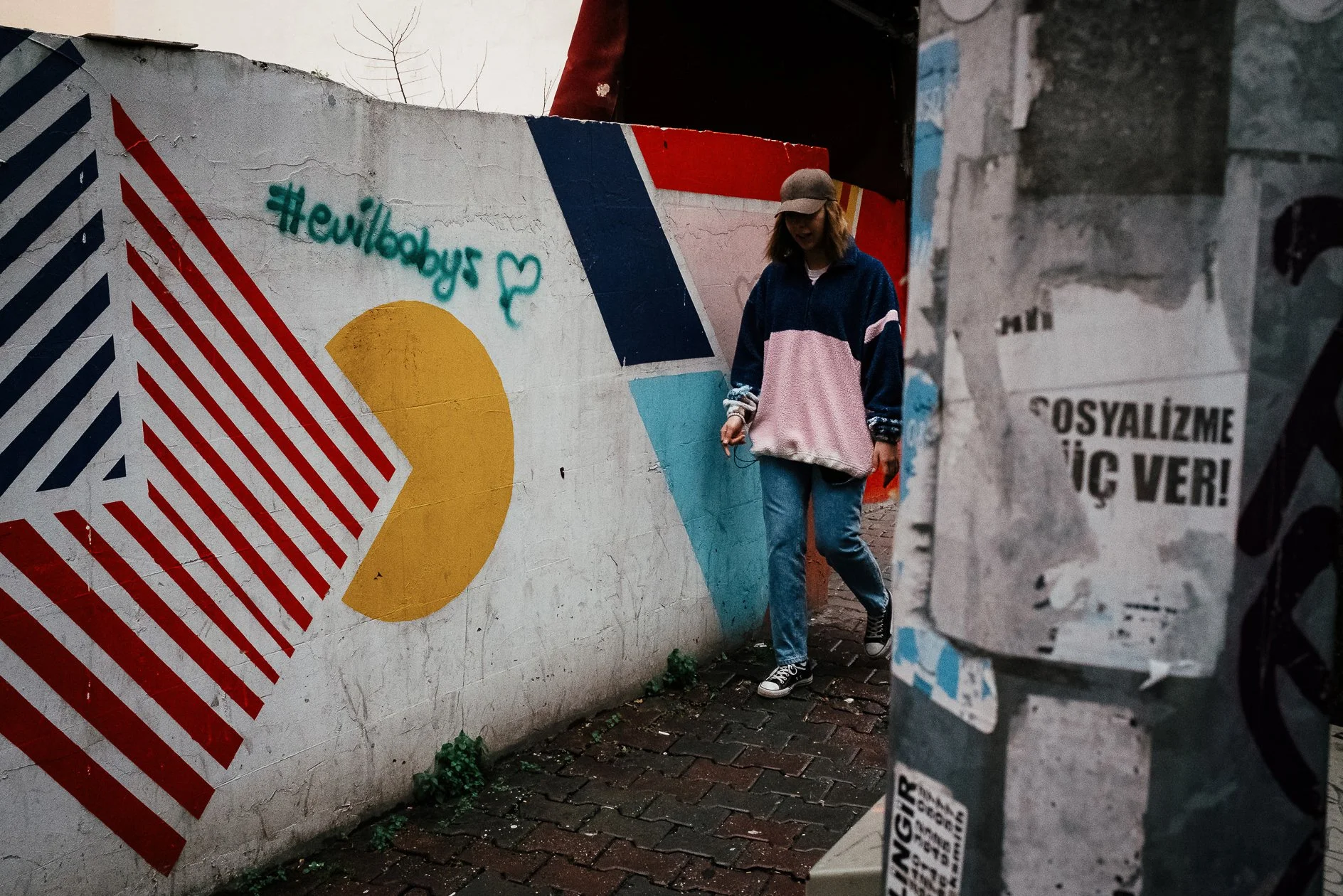 A person walking along a street outside a wall painted with geometric shapes in red, yellow, black, and blue. The wall features a hashtag '#evcilobuy' and a heart symbol. The person is wearing a hat, a color-blocked jacket with pink, dark blue, and w