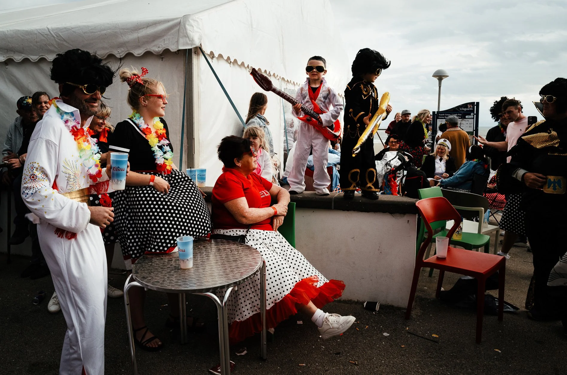People dressed in colorful, retro costumes with wigs and sunglasses gather around a white tent on a boardwalk, some sitting and some standing. Two children are on a small stage holding inflatable guitars, dressed in themed costumes with sunglasses. T