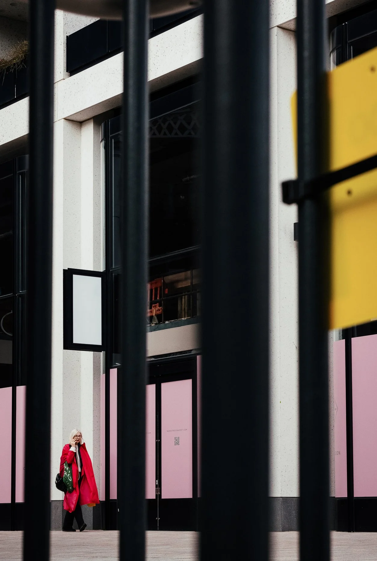 A person in a red coat and glasses walking with a green bag on a city sidewalk, viewed through black metal fences, with a building having pink and black panels in the background.