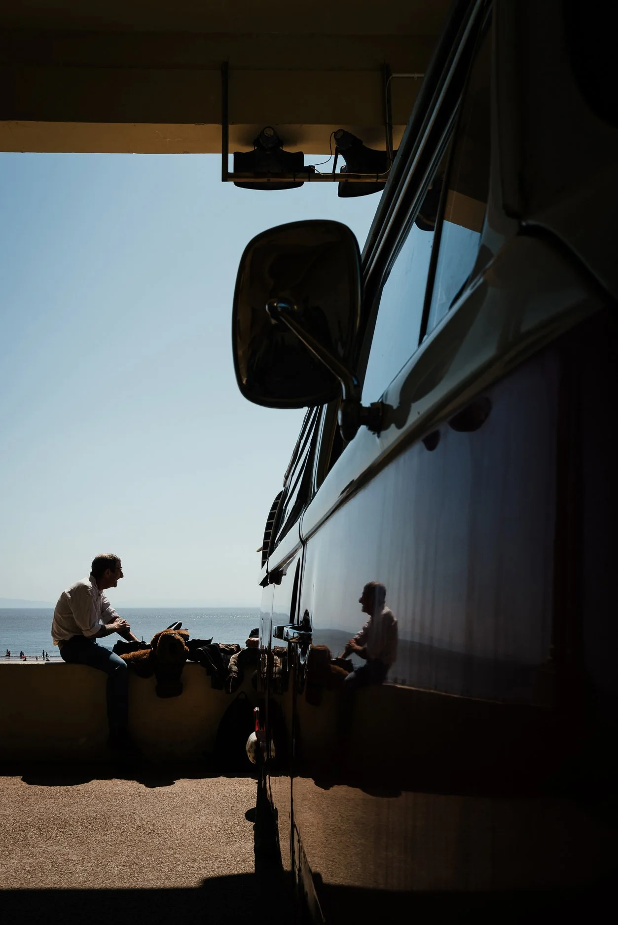 Reflection of people and a car parked by the beach in a shiny, black surface, with the sea and sky in the background. Two people are sitting and one is standing near the car, which is casting a shadow in the foreground.