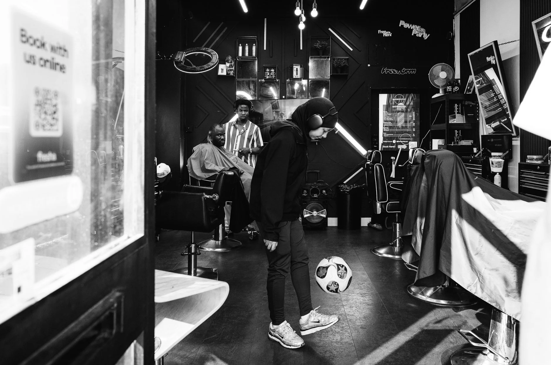 A barbershop with a barber near a customer getting a haircut, two men sitting in chairs, and another person standing with a soccer ball in front of him. The decor features shelves with bottles and neon signs on a dark wall, and there are barber chair